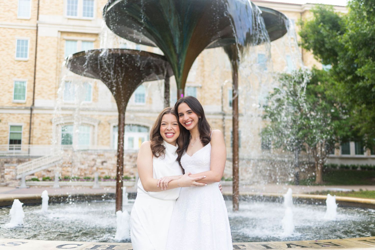 Two women in white dresses smiling and embracing in front of a decorative fountain outdoors.