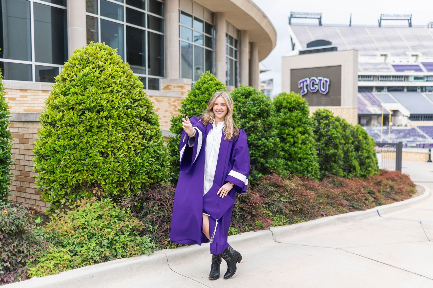 Woman in purple TCU graduation gown posing outside Amon G. Carter Stadium, tossing cap and smiling.