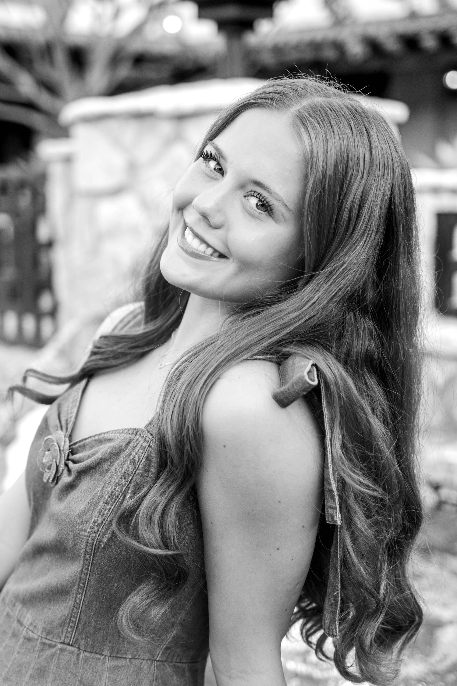 Smiling young woman with long wavy hair wearing denim overalls poses outdoors in a black and white portrait photo.