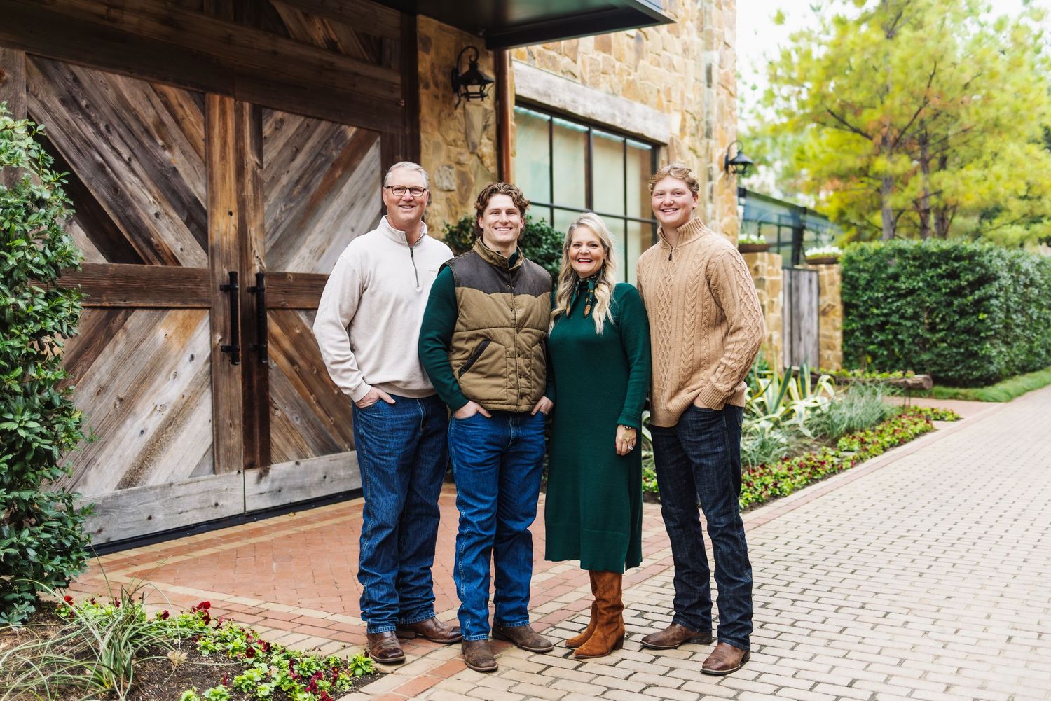 Four smiling people pose together outdoors near rustic barn doors and lush greenery on a cloudy day.