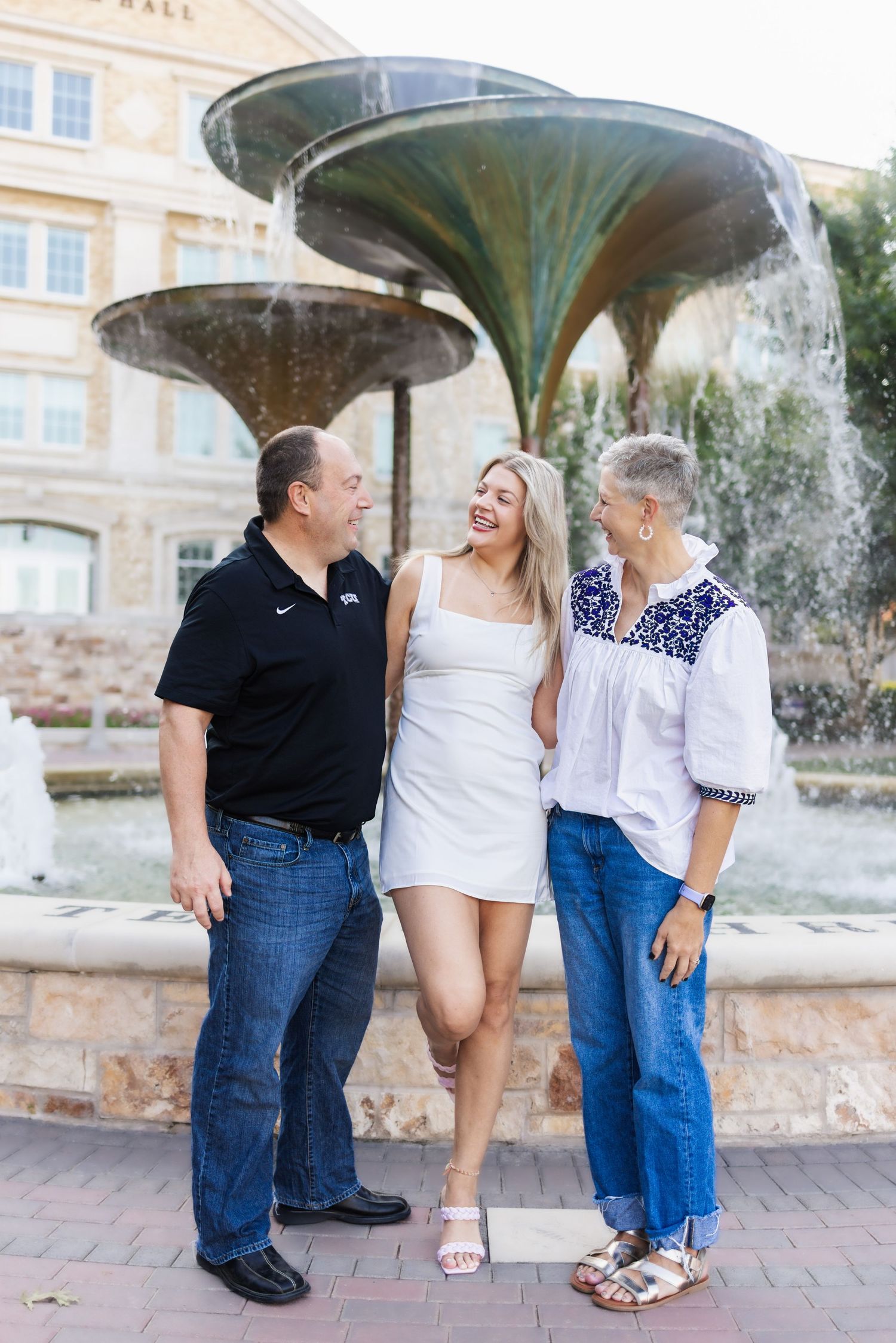 TCU graduate in white dress celebrates with her parents in front of a campus fountain.