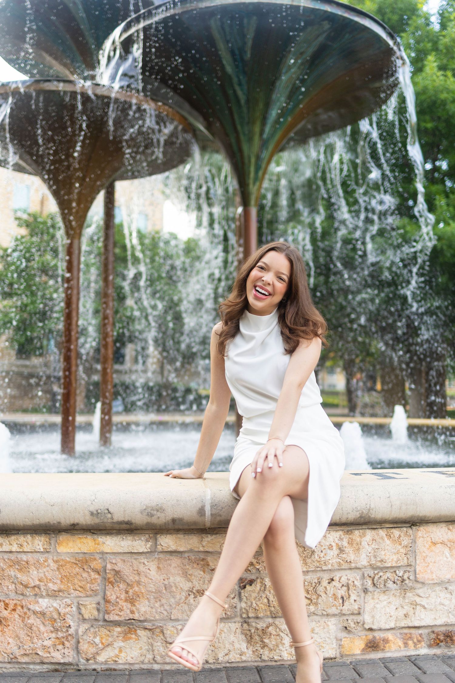 Young woman in white dress laughing by ornate fountain outdoors.