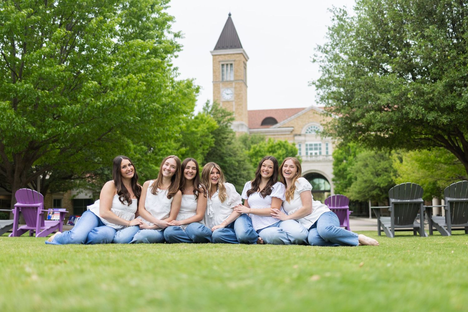 Seven young women in white tops and jeans sitting together on green campus lawn with clock tower building behind them.