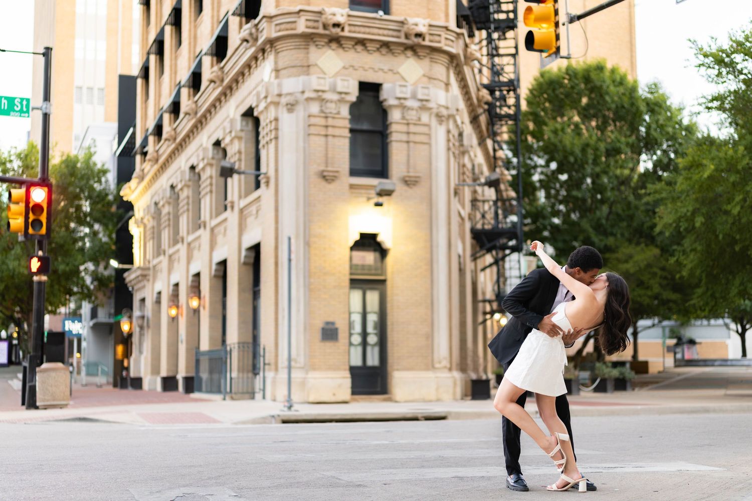 Couple sharing a romantic dip kiss on a city street corner with a historic beige building in the background.