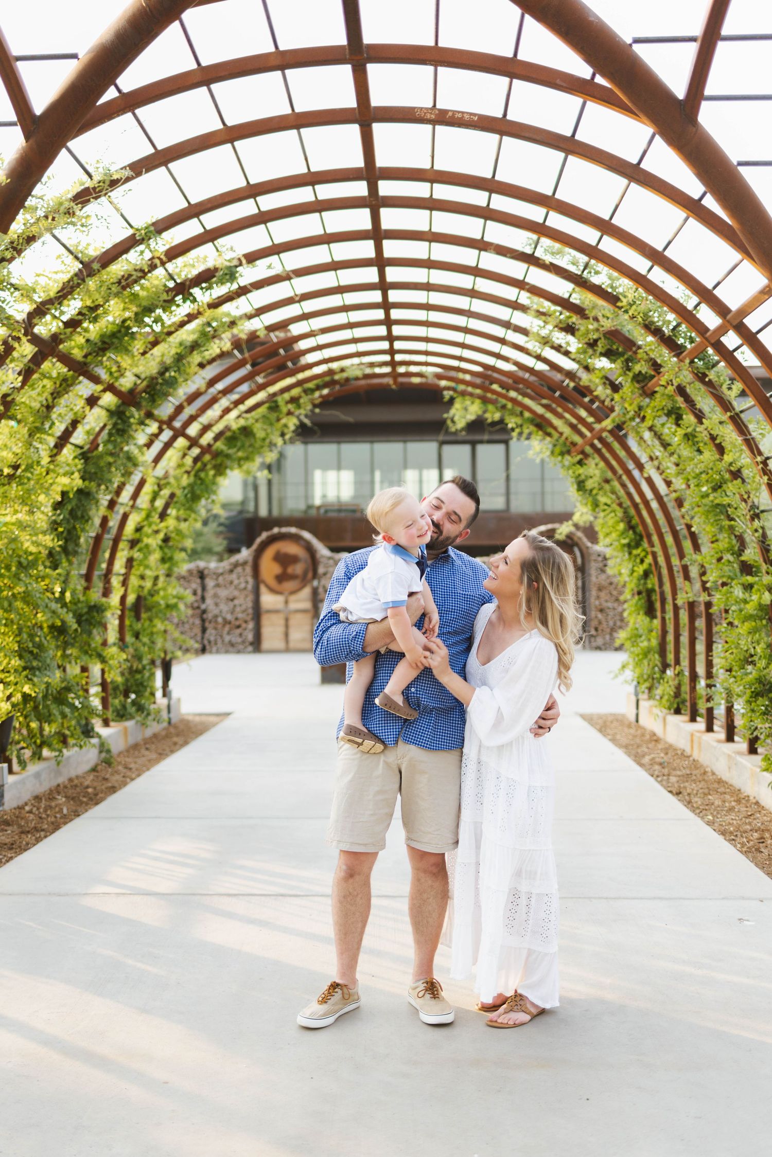 Family of three posing under a rustic arched pergola with lush greenery, parents holding toddler son outdoors.