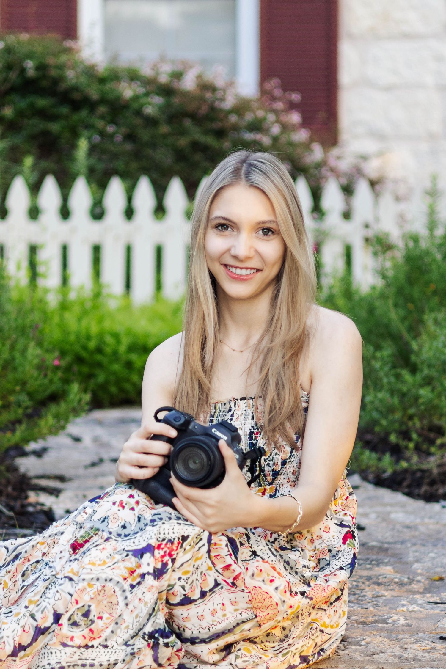 Smiling blonde woman in floral dress sitting outdoors holding a DSLR camera near a white picket fence.