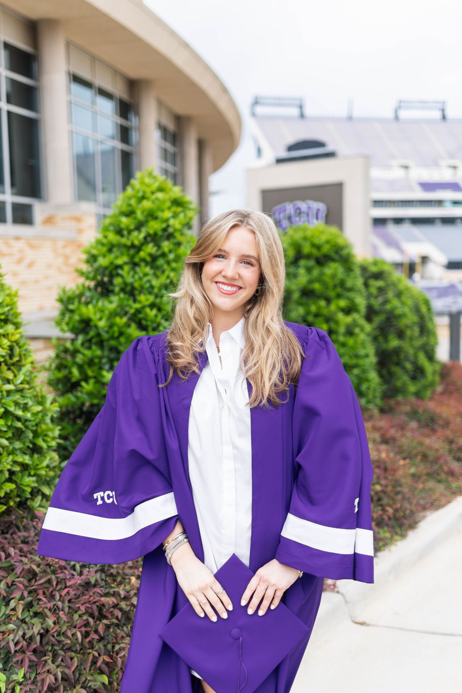 Smiling blonde woman in purple TCU graduation gown poses outdoors near stadium.
