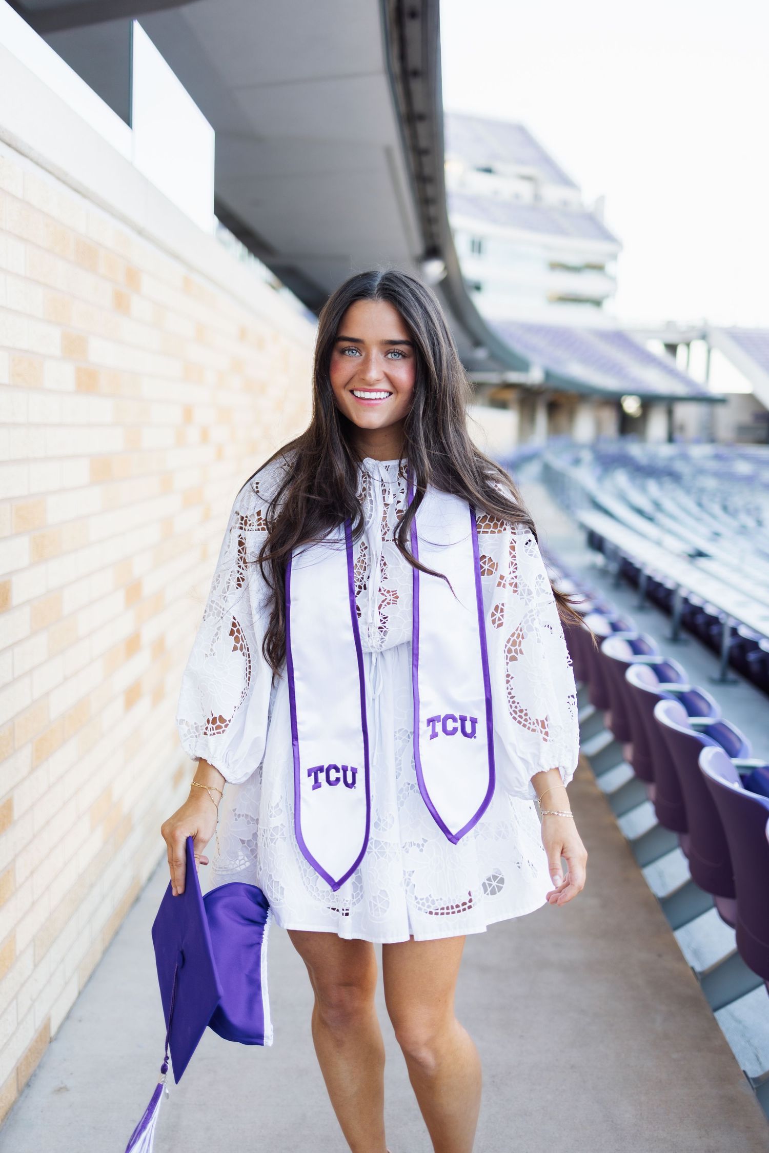 Happy TCU graduate in white dress and purple stole walking through stadium corridor holding cap.