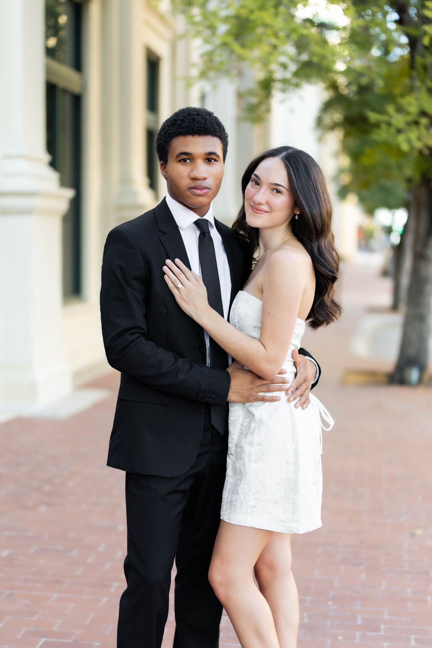 Elegant interracial couple posing outdoors, man in black suit with tie, woman in white mini dress, urban backdrop.