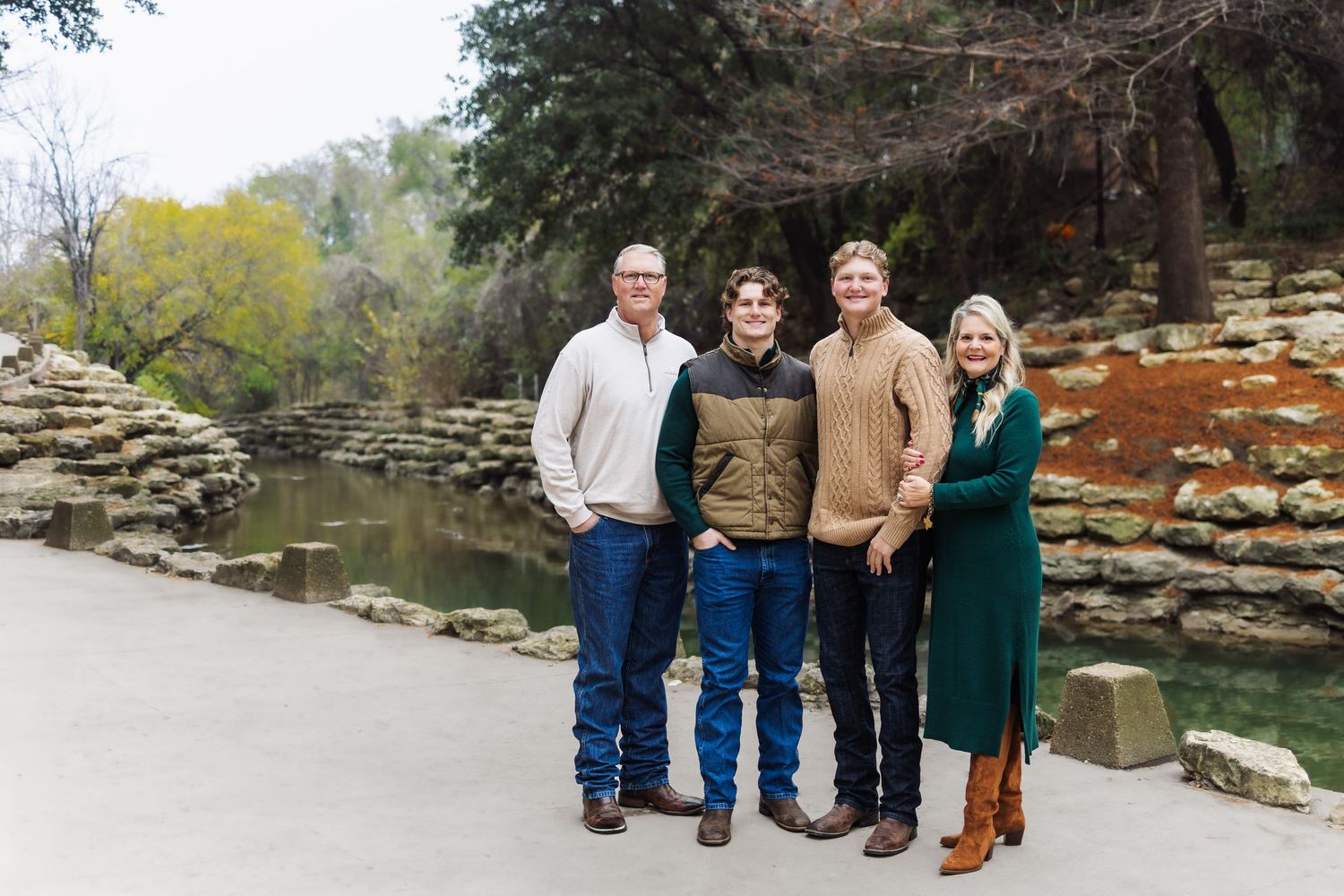 Family of four smiling outdoors near a rocky creek, dressed in casual fall attire, surrounded by lush greenery.