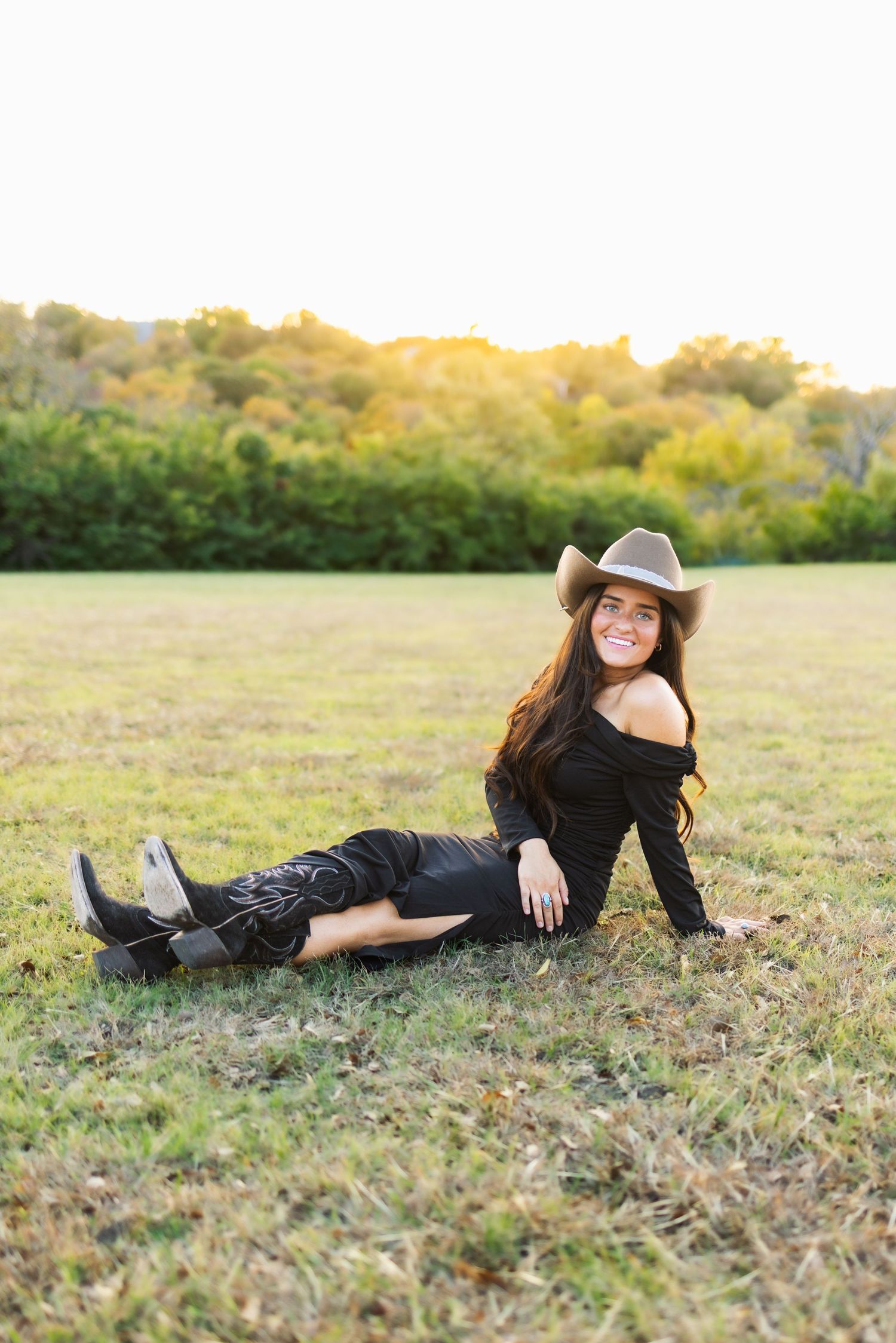 Young woman in black outfit, cowboy hat, and boots sitting on grass field at golden hour sunset.