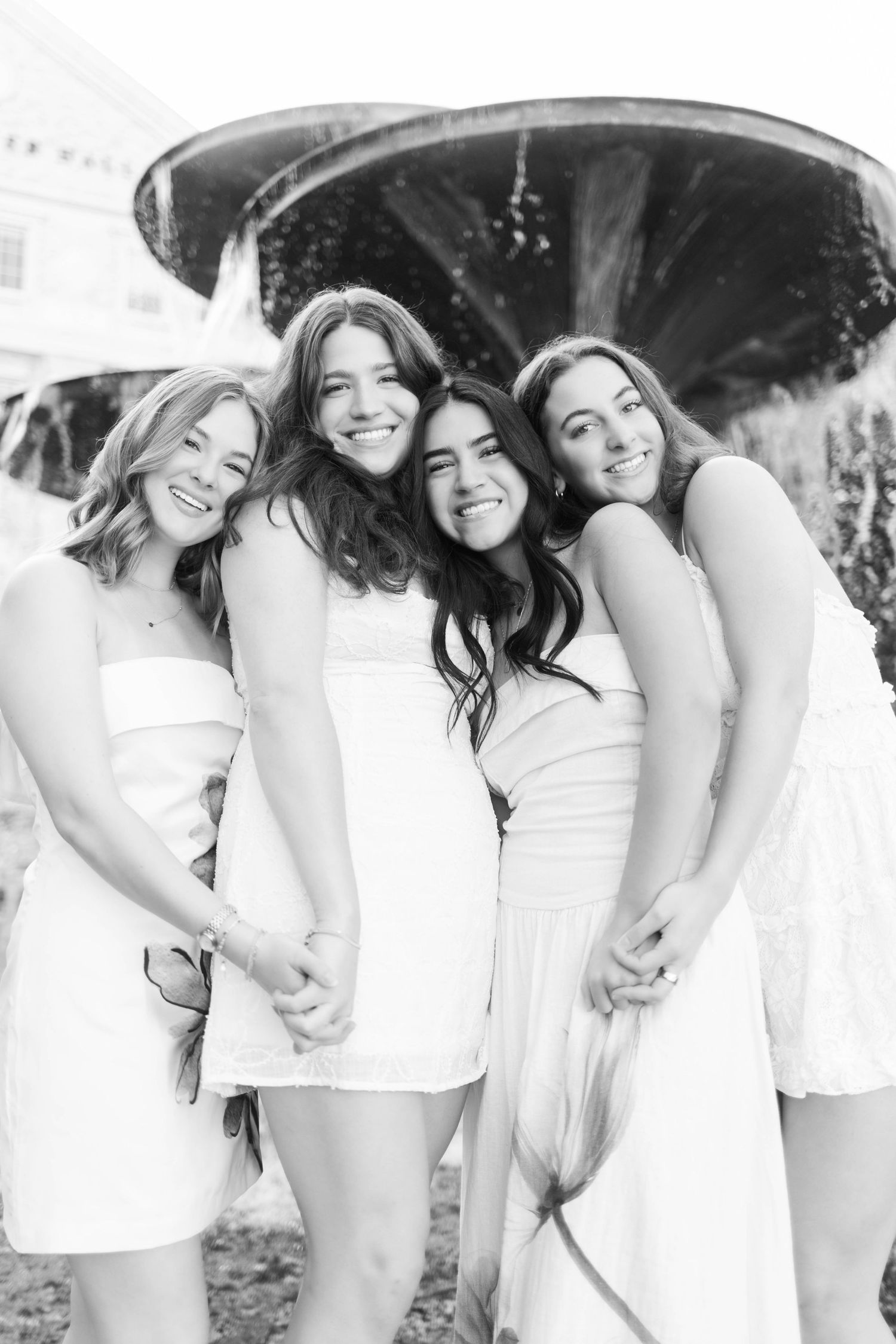 Black and white photo of four young women in white dresses posing together by a decorative fountain.