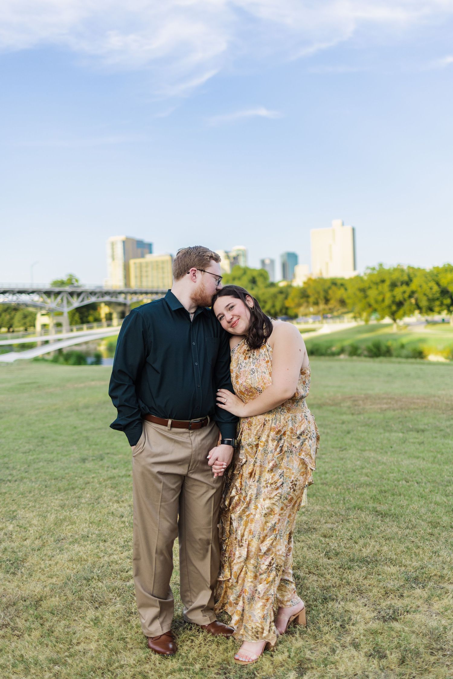 Couple sharing a romantic moment on a green lawn with a city skyline backdrop on a sunny day.