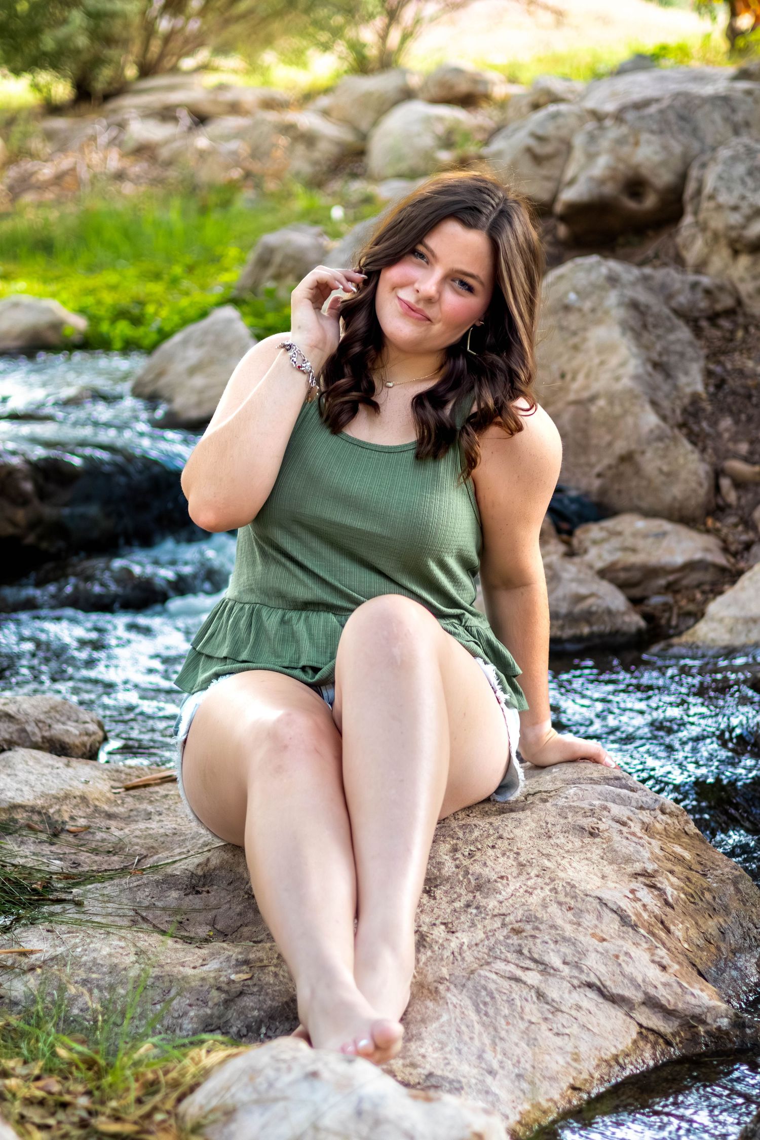 Young woman in green dress sitting barefoot on rocks by a flowing stream in a lush outdoor setting.