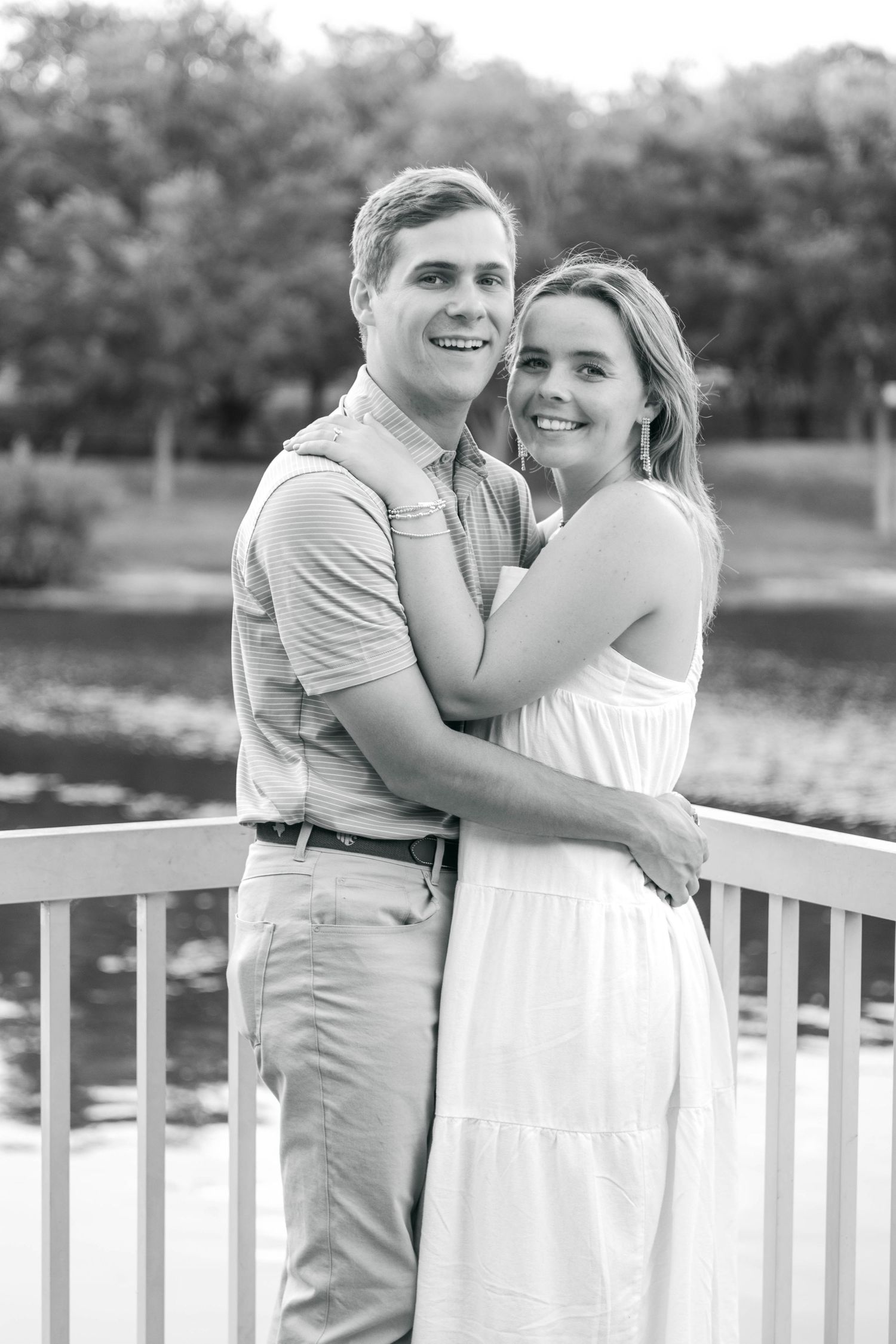 Black and white photo of a smiling couple embracing on a bridge near a pond.