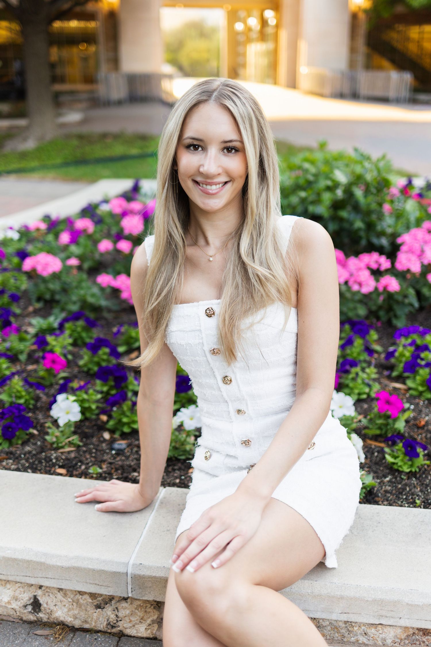 Young blonde woman in white dress sitting by colorful flower garden, smiling outdoors.