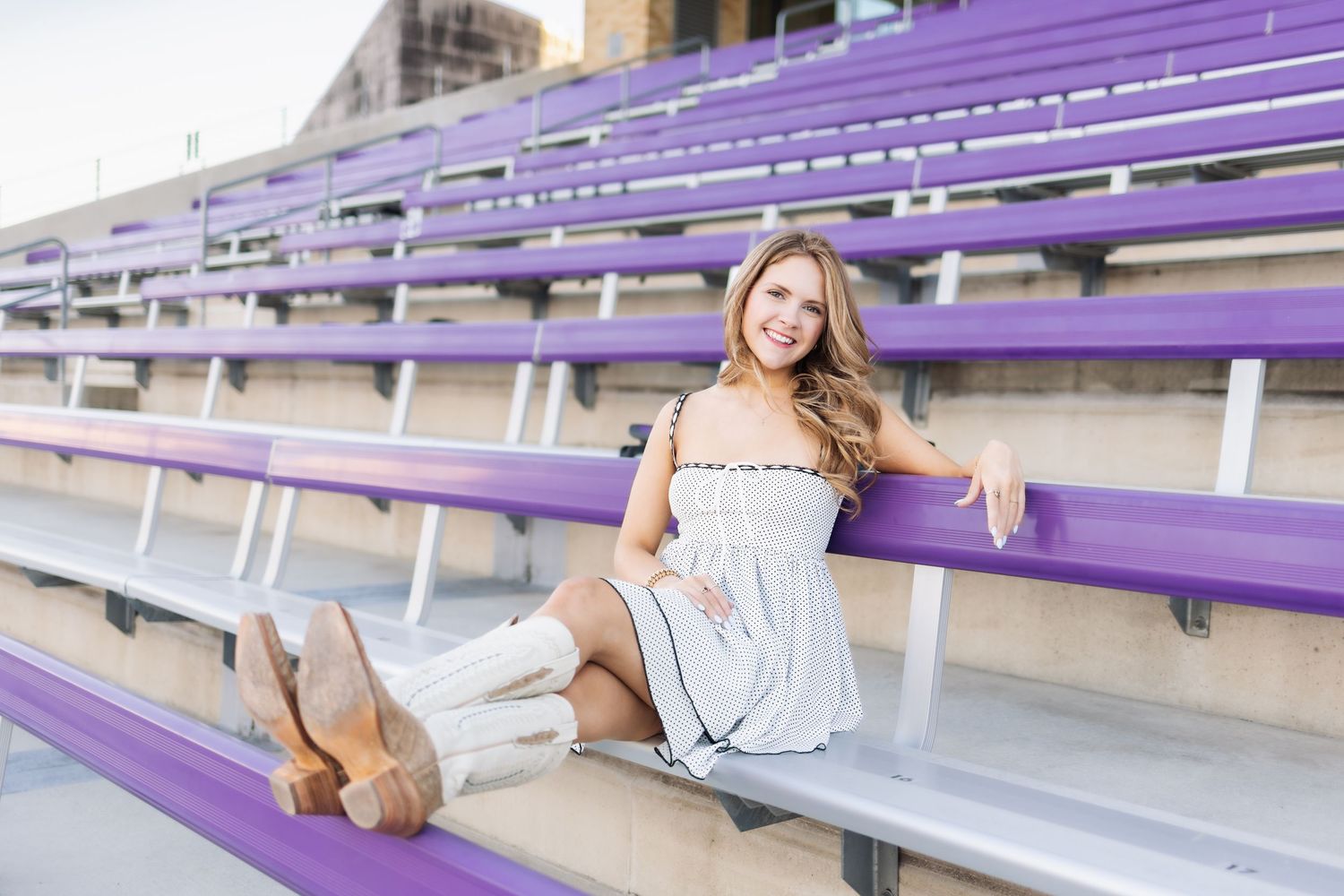 Young woman in white dress and boots smiling on purple bleachers at a stadium.