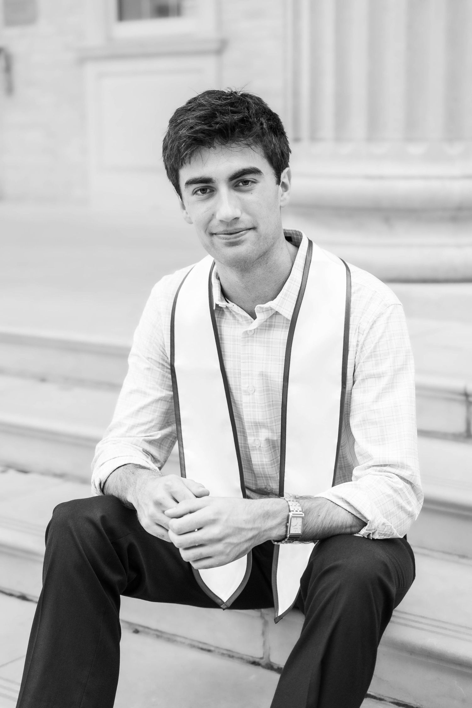 Young man in white shirt and graduation stole sits on steps in black and white photo.
