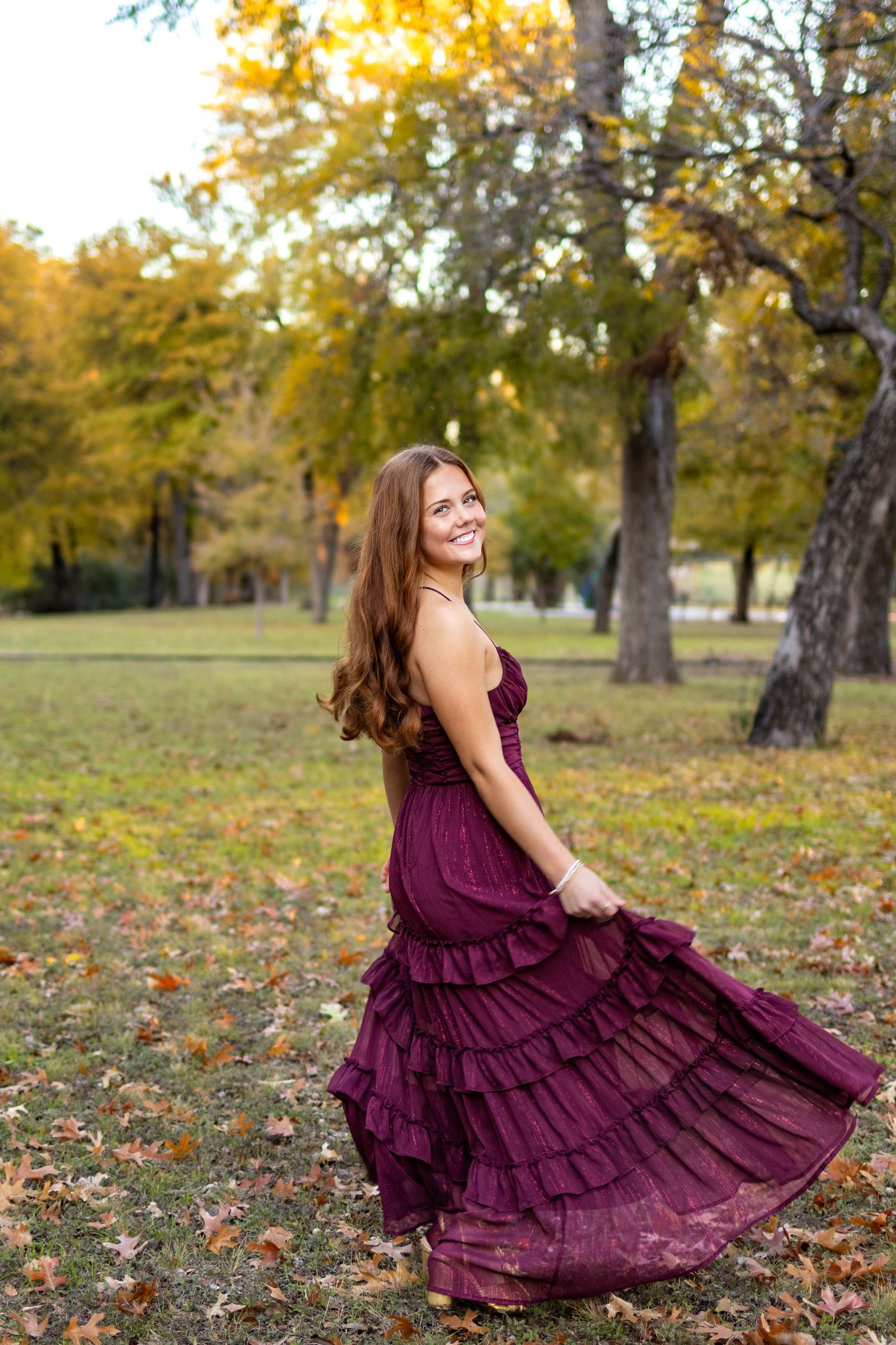 Young woman in burgundy ruffled dress twirling in autumn park, smiling over shoulder, fall foliage background.