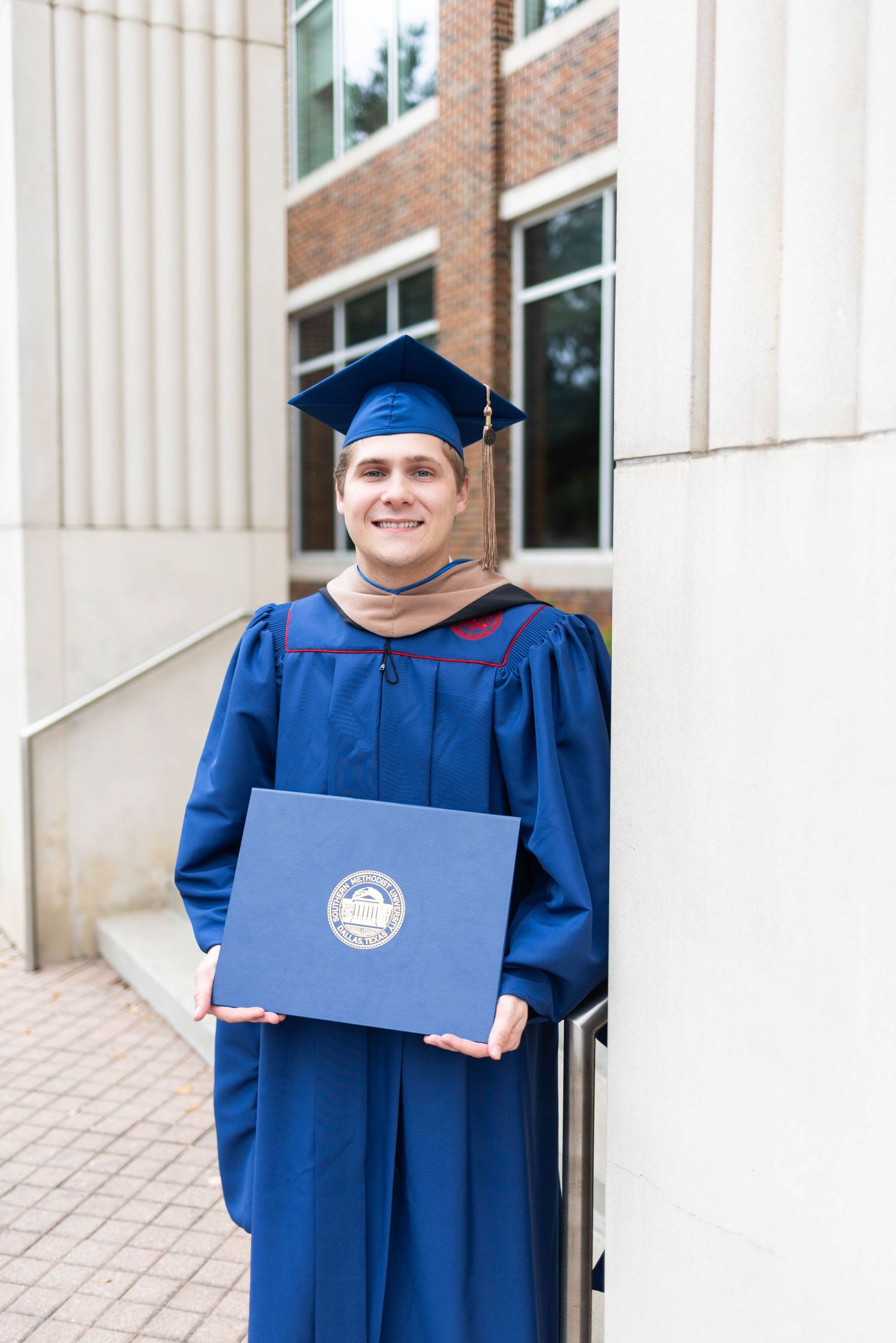 Smiling male graduate in blue cap and gown holds diploma cover outside a university building.