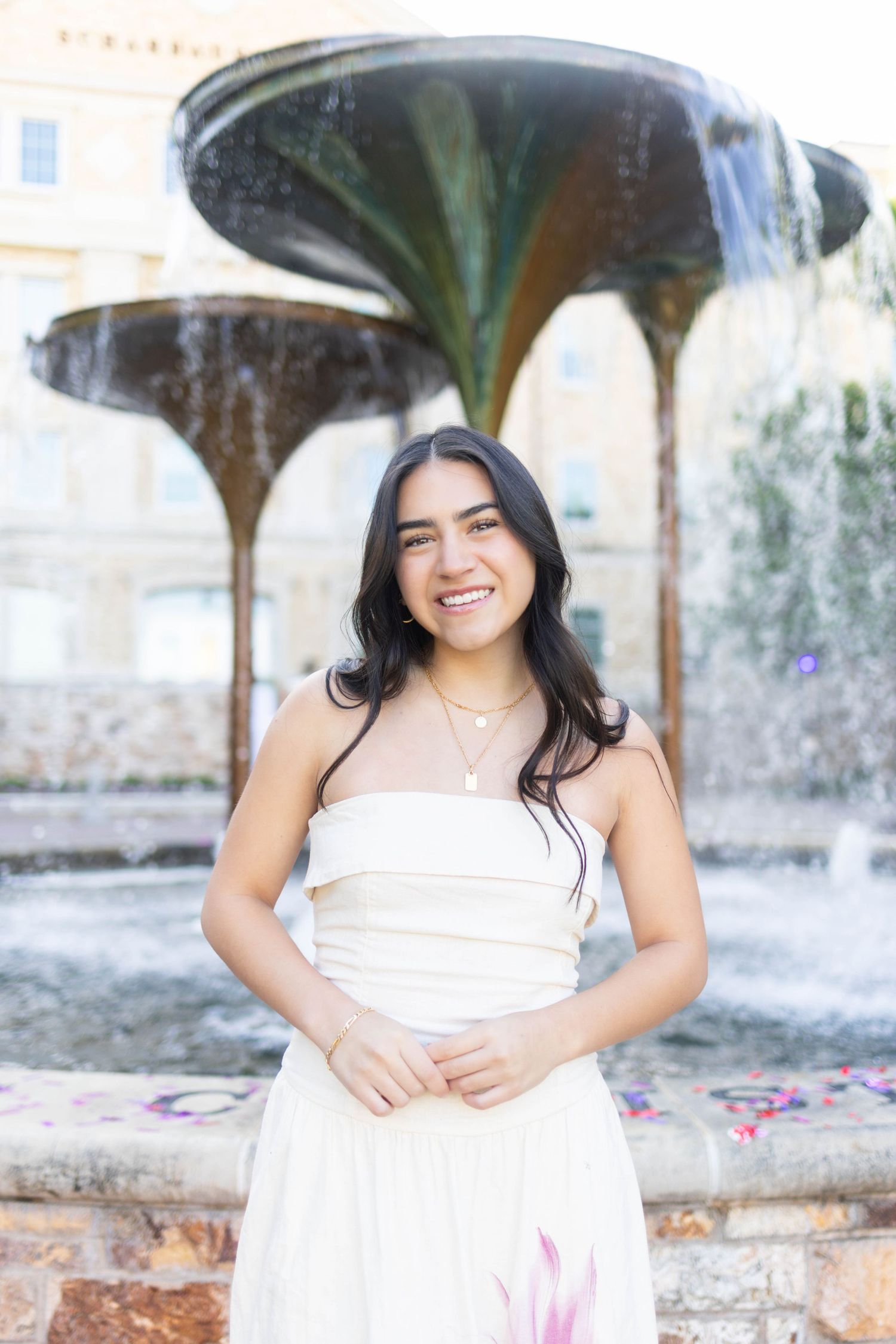 Young woman in white strapless dress smiling in front of ornate fountain outdoors.