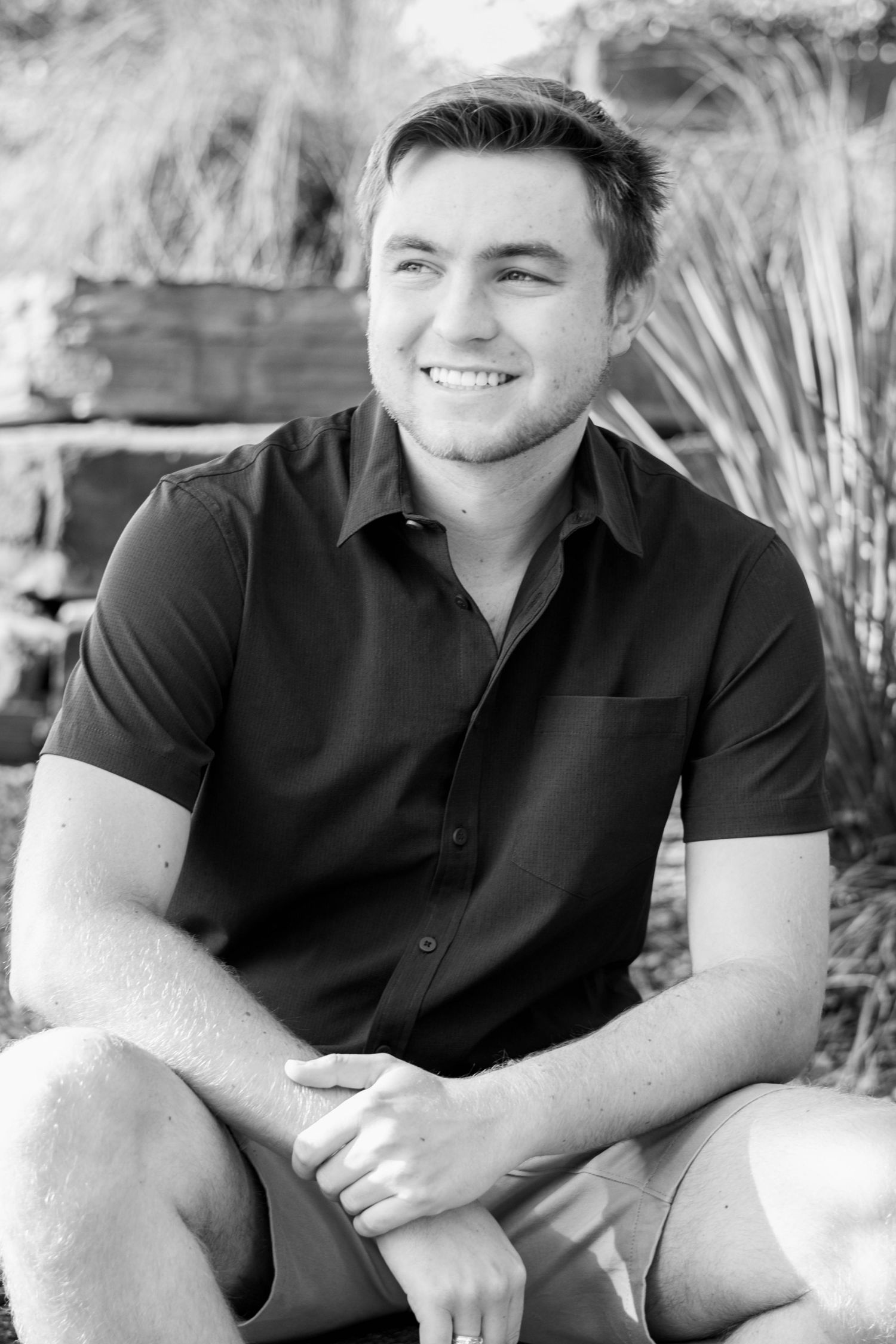 Black and white portrait of a smiling young man in a dark shirt sitting outdoors near tropical plants.