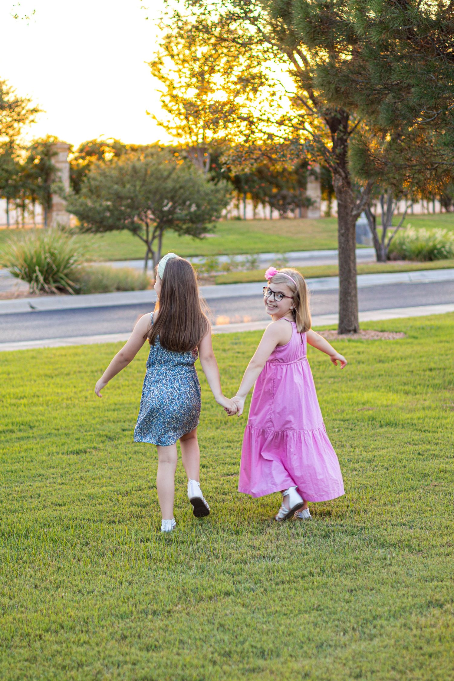 Two young girls holding hands and walking on green grass at sunset, one in pink dress turning to smile at camera.