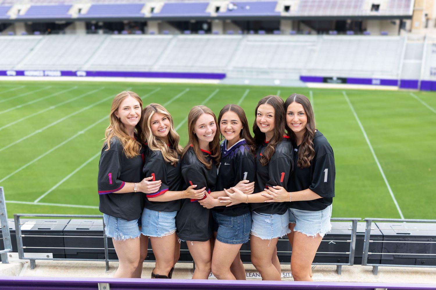 Six young women in matching black jackets and denim shorts pose smiling at a purple college football stadium.