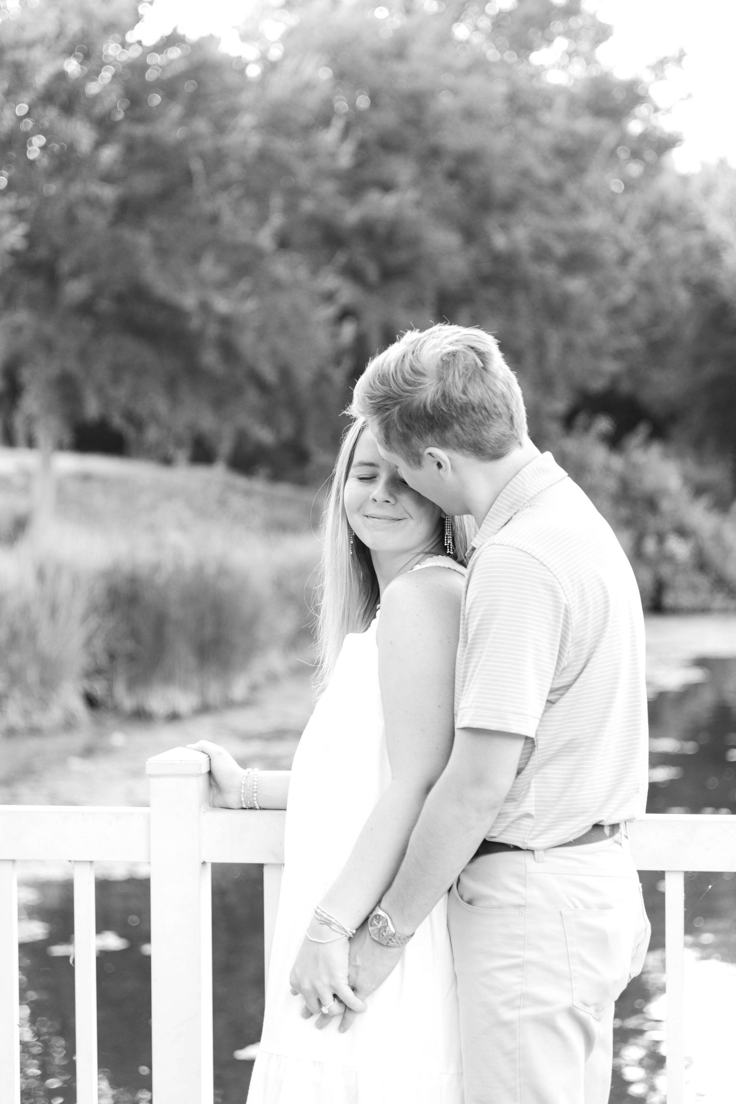 Black and white photo of a couple holding hands on a bridge, man kissing womans head.