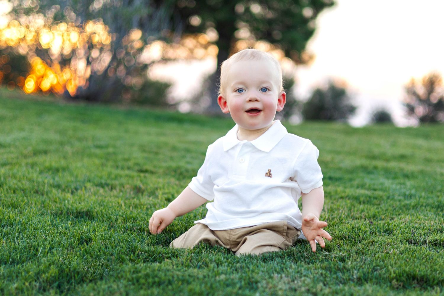 A happy baby boy in a white polo shirt sits on lush green grass outdoors during golden hour.