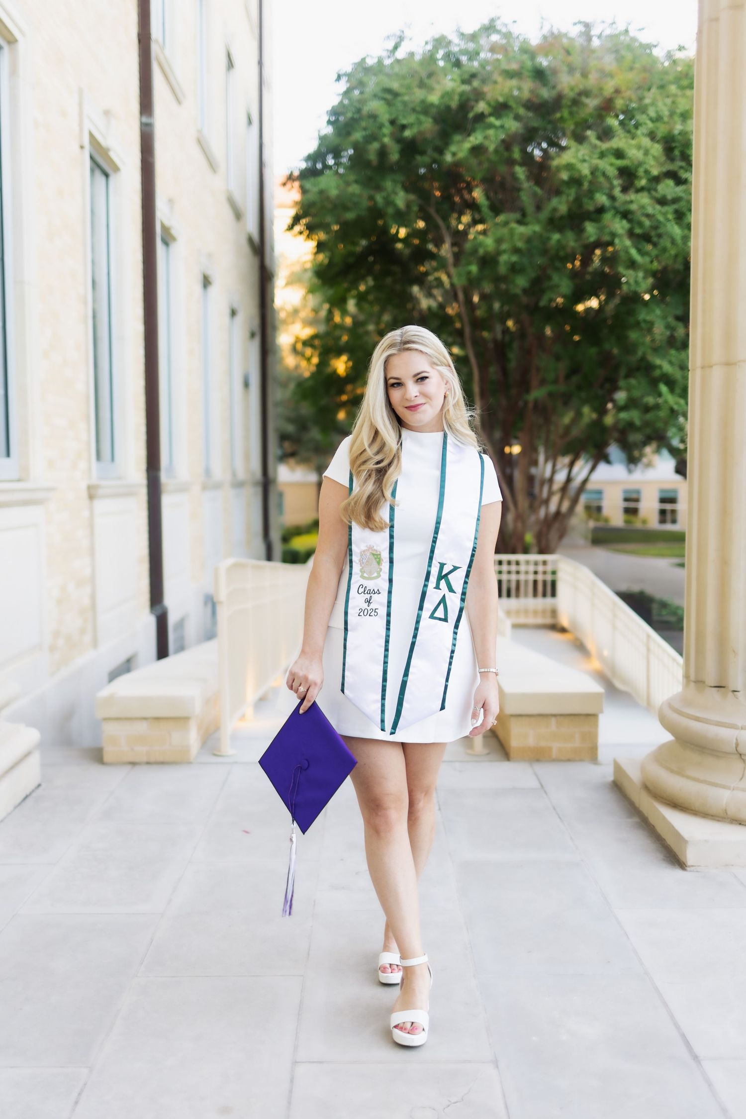 Smiling blonde woman in white dress holding purple graduation cap wearing Kappa Delta stole.
