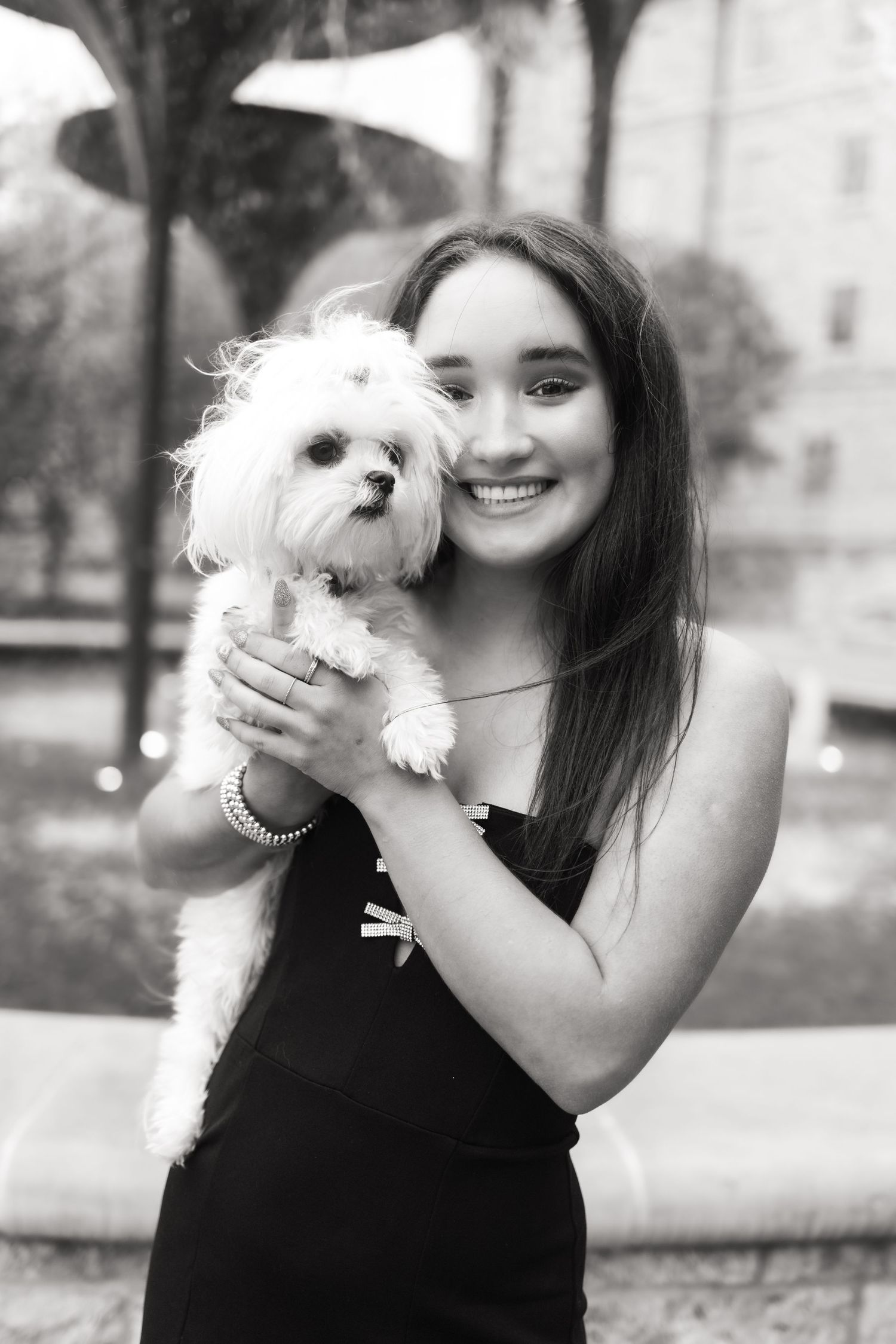 Black and white portrait of a smiling young woman holding a small white Maltese dog near a fountain.