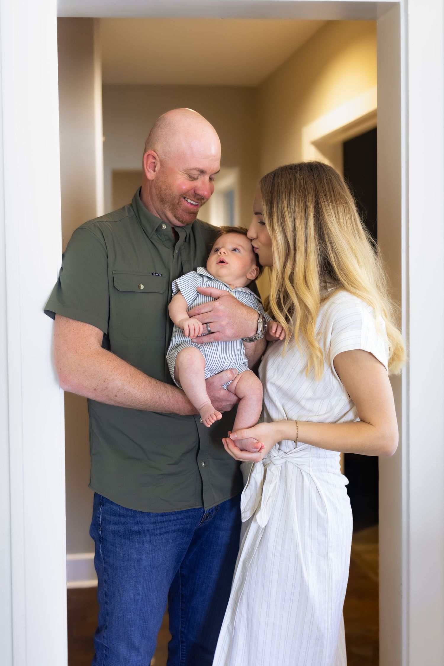 Happy couple holding their newborn baby, smiling and sharing a tender kiss in a bright home hallway.