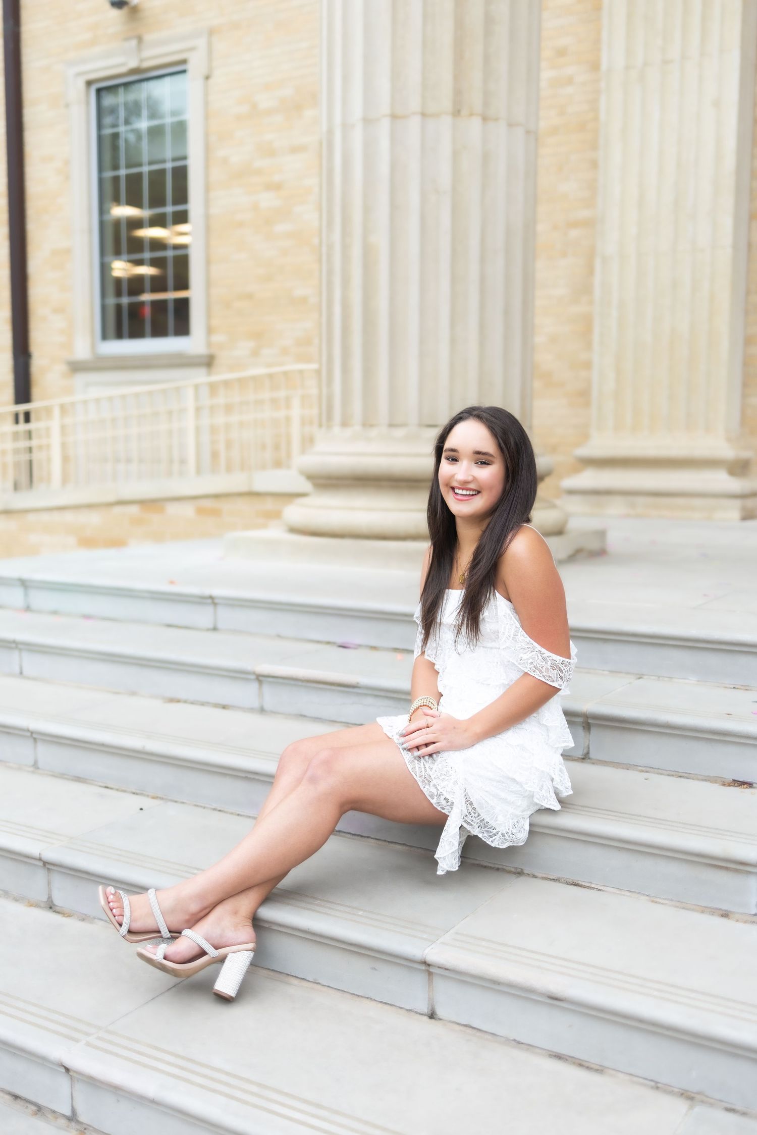 Young woman in white off-shoulder dress and heels sitting on stone steps outside a classical building.