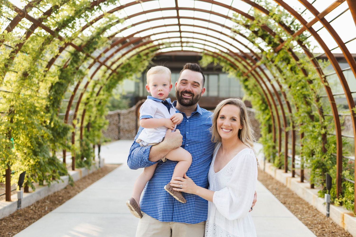 A smiling family of three poses under a lush green garden archway, with dad holding a baby boy.