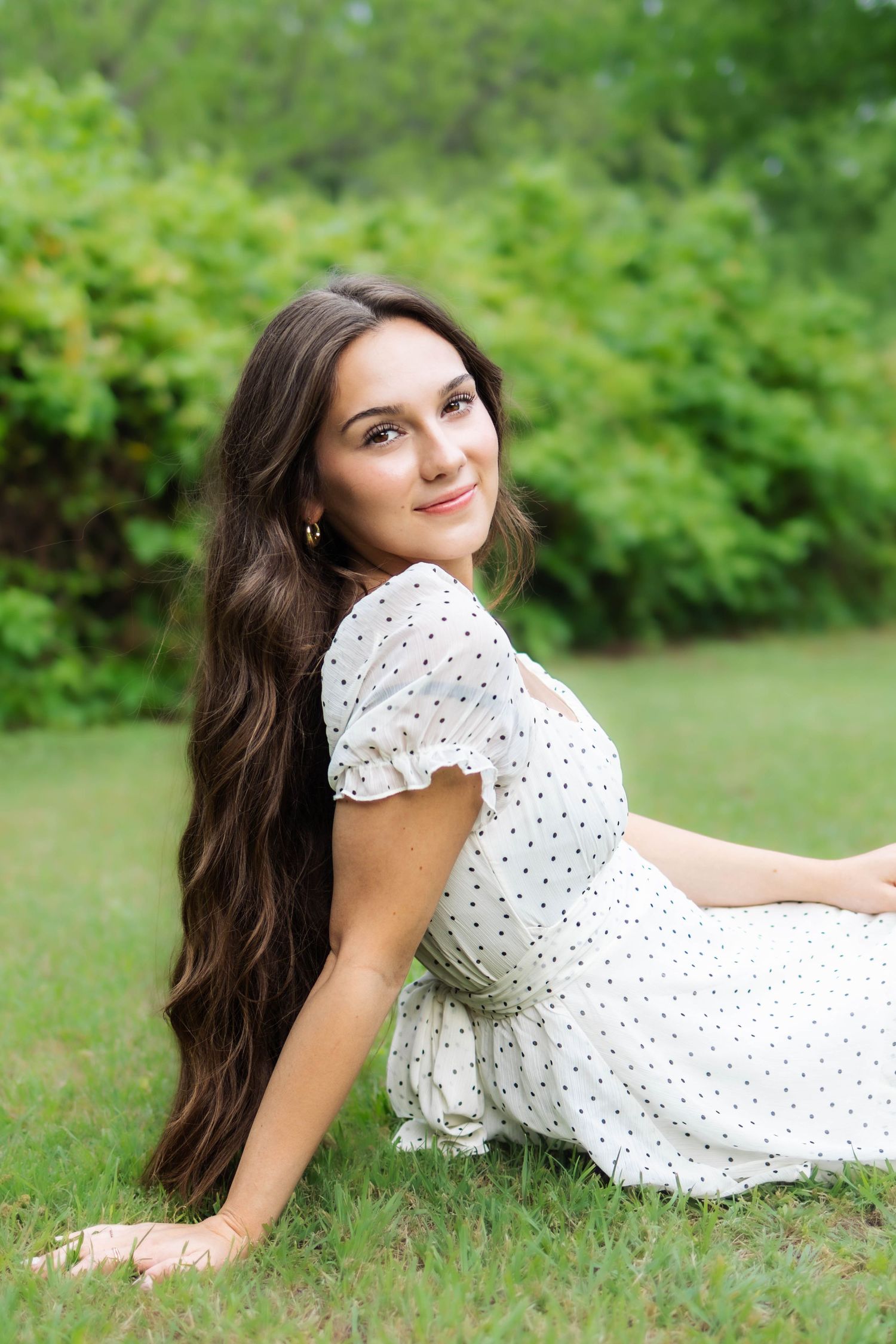 Brunette woman in white polka dot dress sitting on green grass with trees behind her.