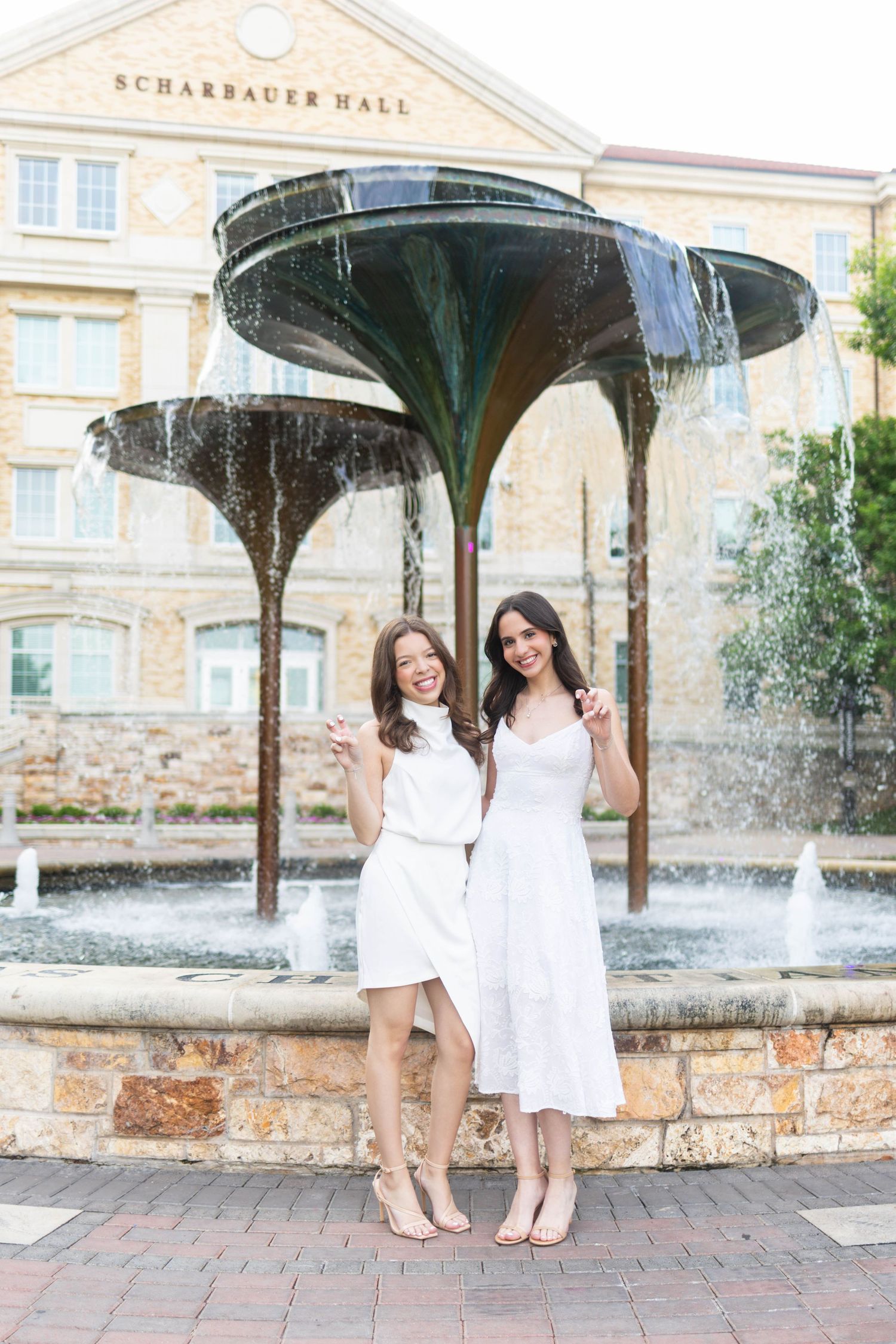 Two women in white dresses pose smiling by a fountain outside Scharbauer Hall on a sunny day.