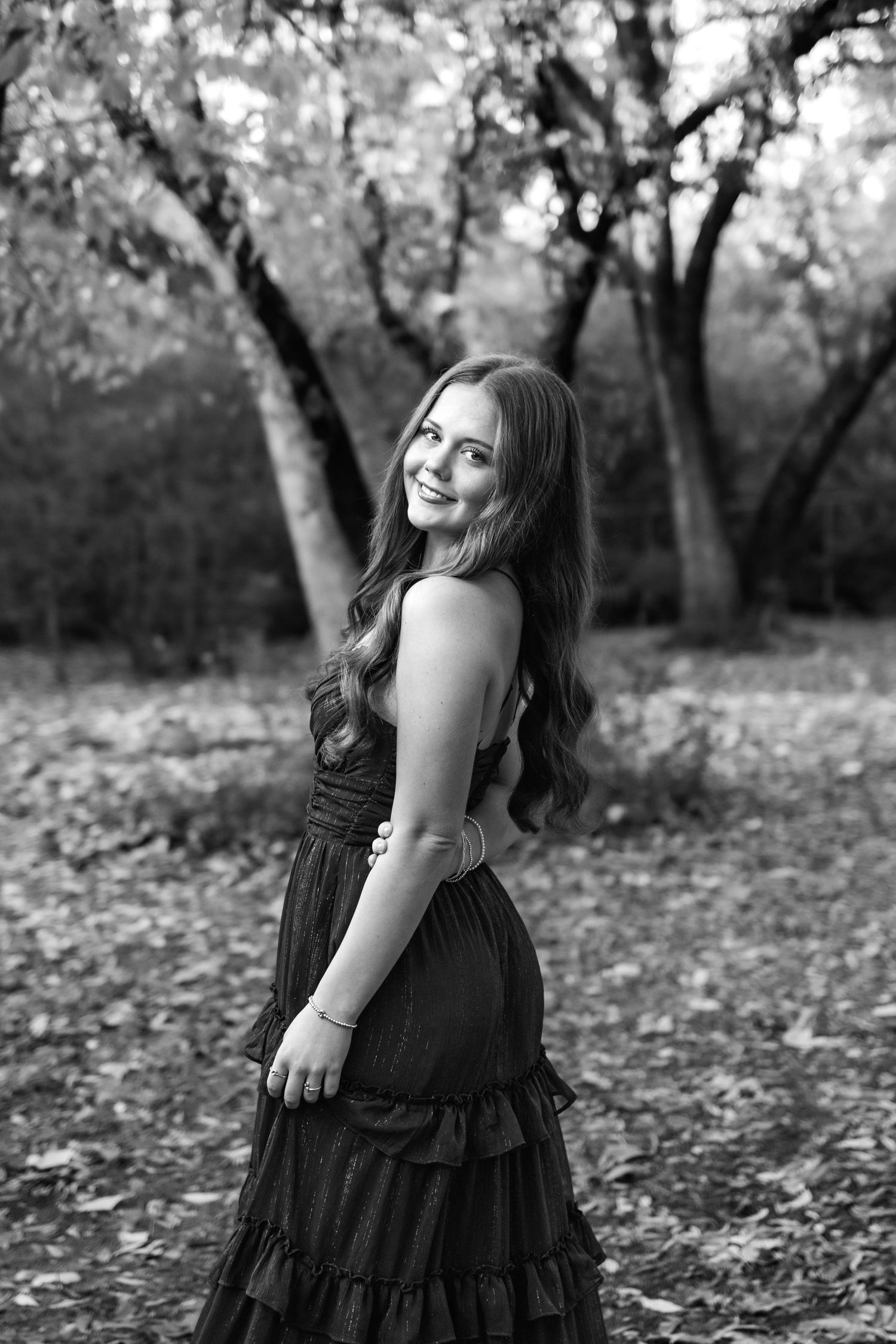 Black and white portrait of a smiling young woman in a ruffled dress posing outdoors among autumn trees.