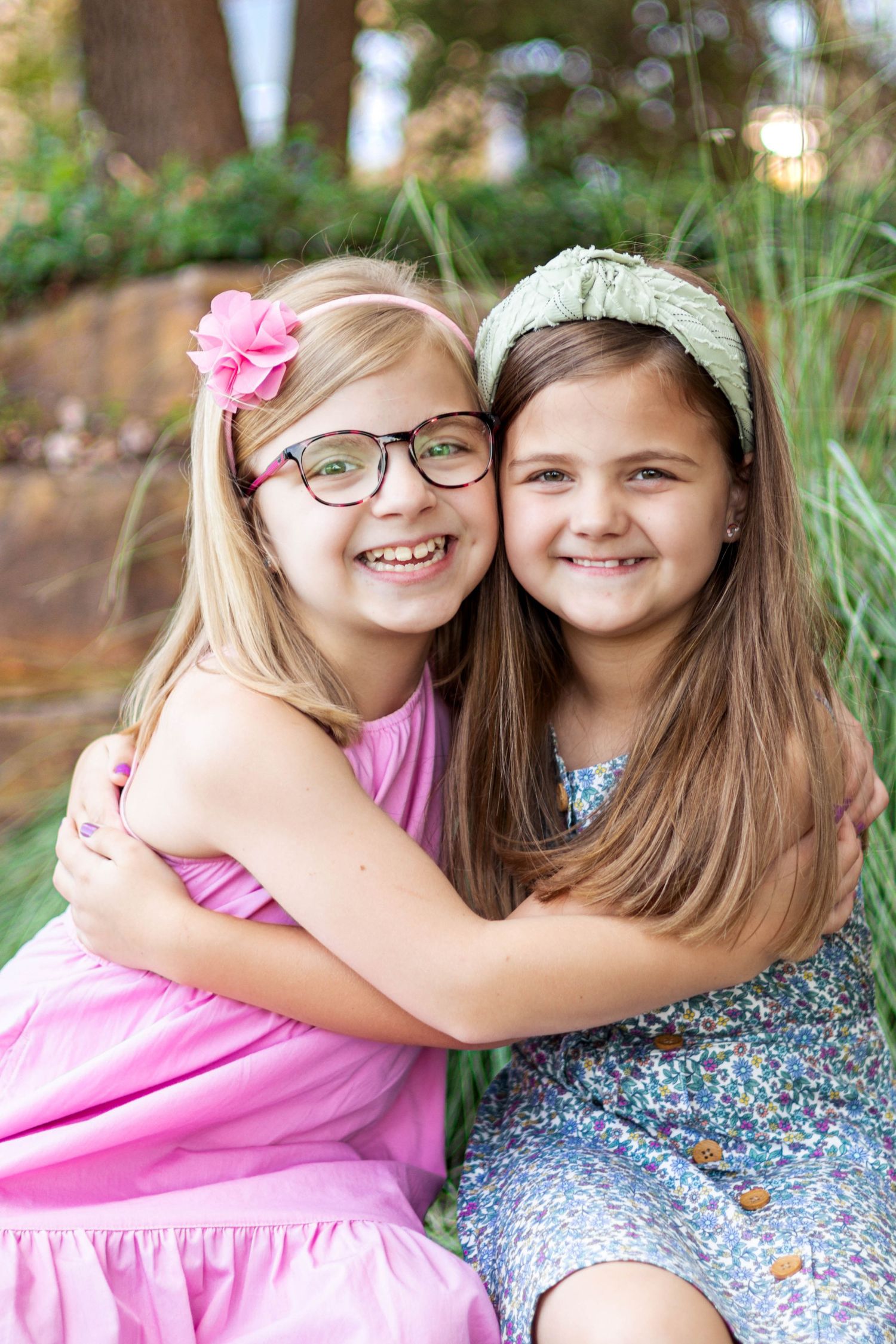 Two young girls hugging and smiling outdoors, one wearing pink dress and glasses, other in floral dress with green headband.