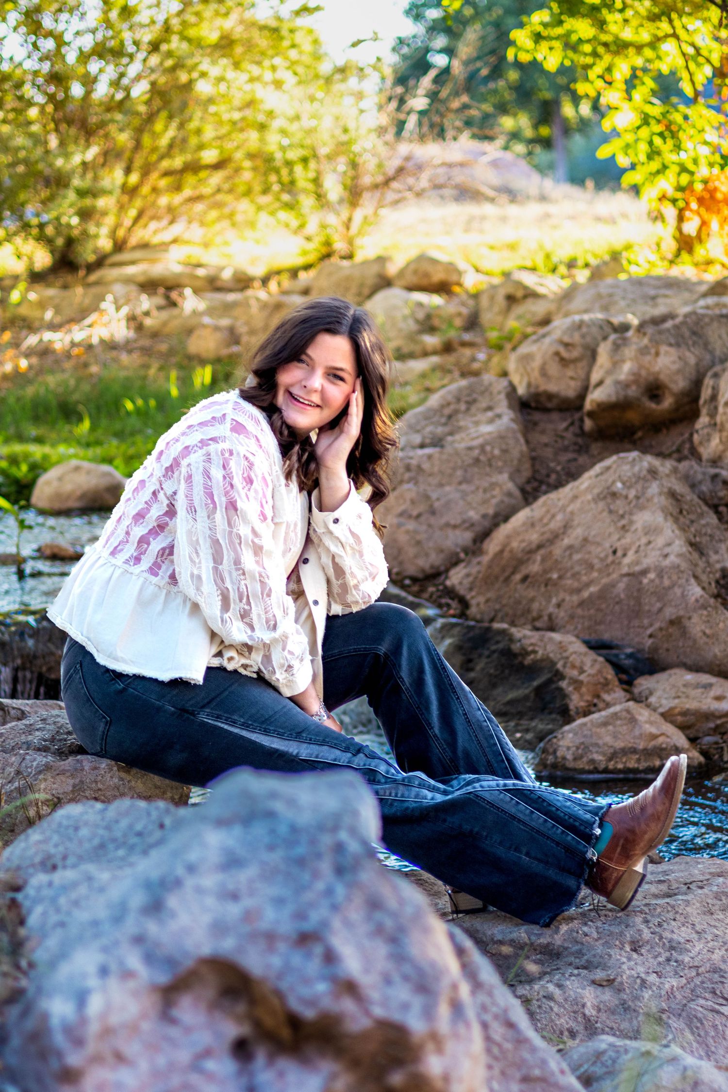 A smiling woman with dark hair sits on rocks near a stream, wearing a floral blouse, dark jeans, and brown boots.