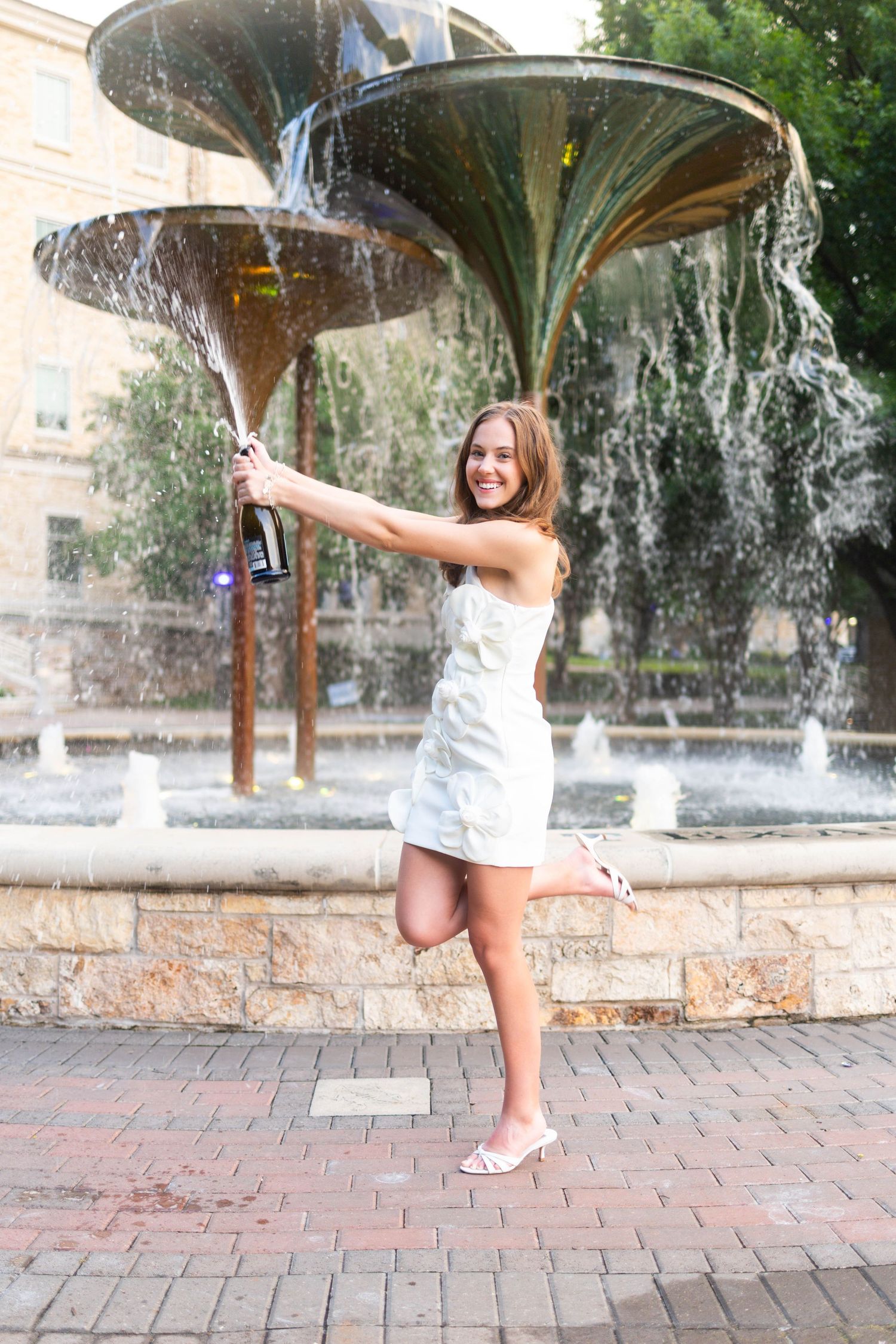 Smiling woman in white dress pops champagne bottle near outdoor fountain in celebration.