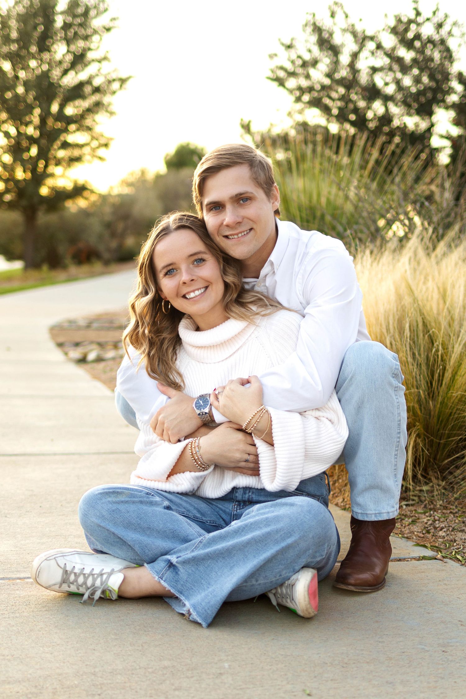 A smiling couple in white sweaters and jeans poses outdoors on a pathway, with the man hugging the woman from behind.