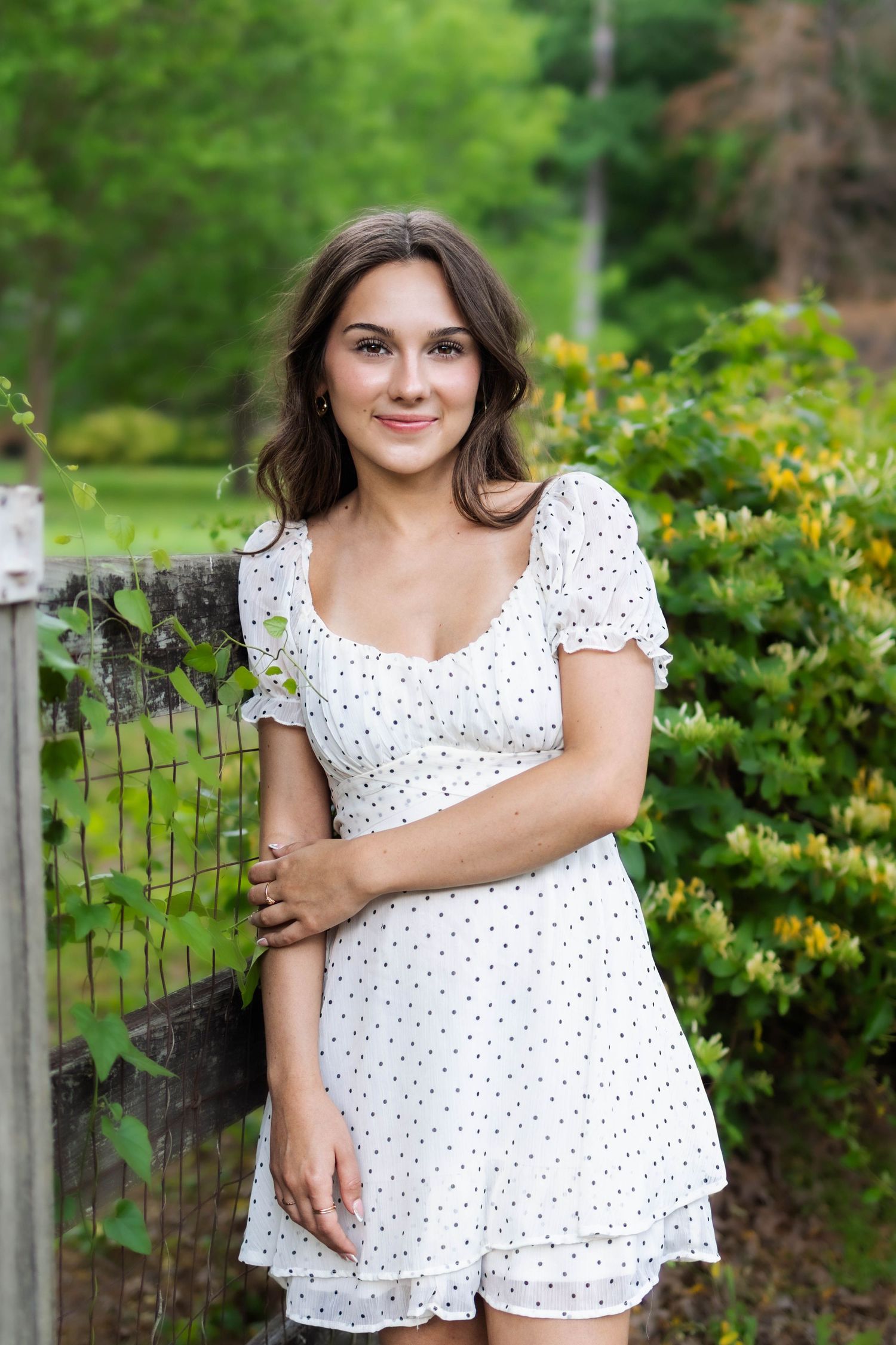 Young woman in white polka dot dress posing outdoors near a wooden fence with lush green bushes.