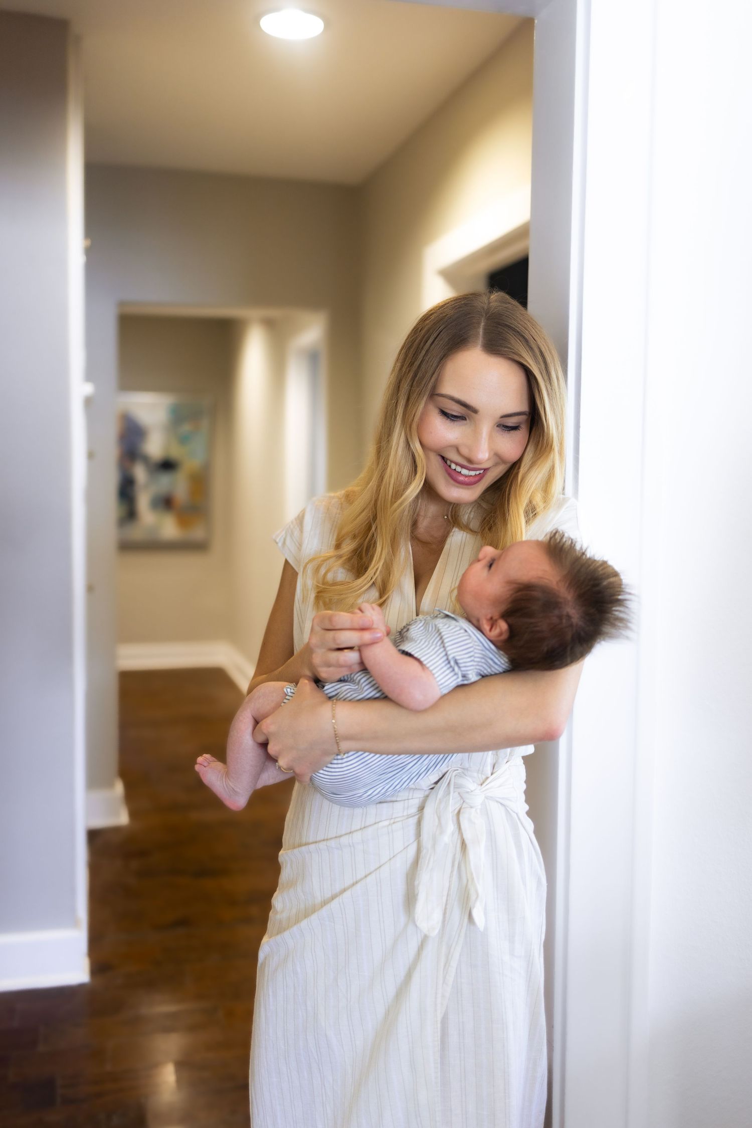 Smiling young mother holding her newborn baby in a bright home hallway, wearing a striped dress.