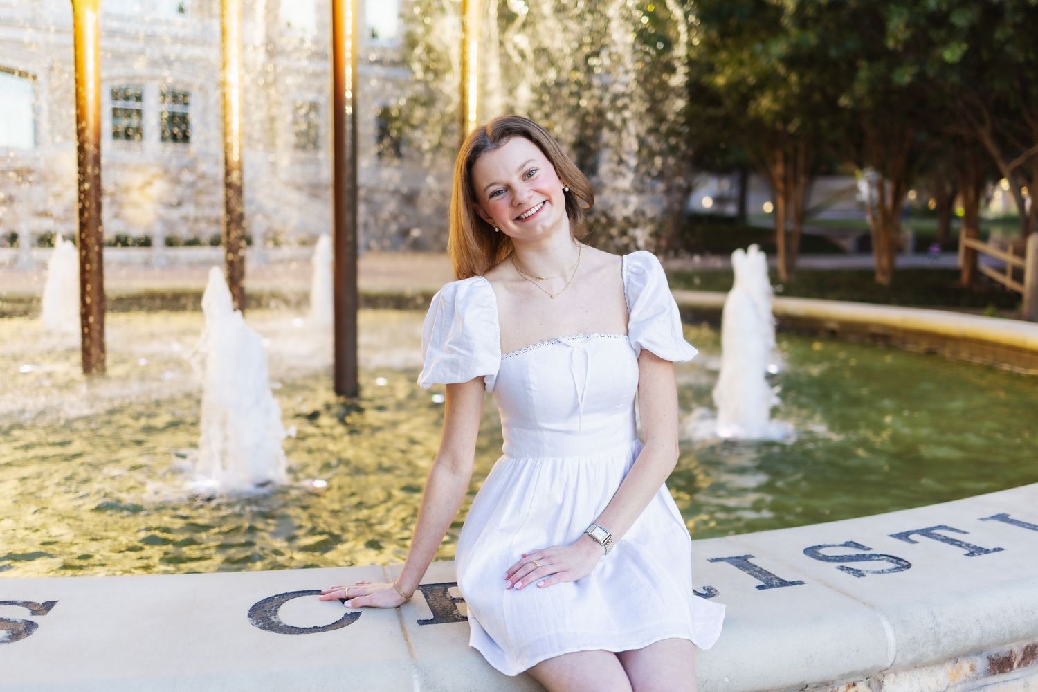 Young woman in white dress smiling by a fountain in a sunny outdoor plaza.