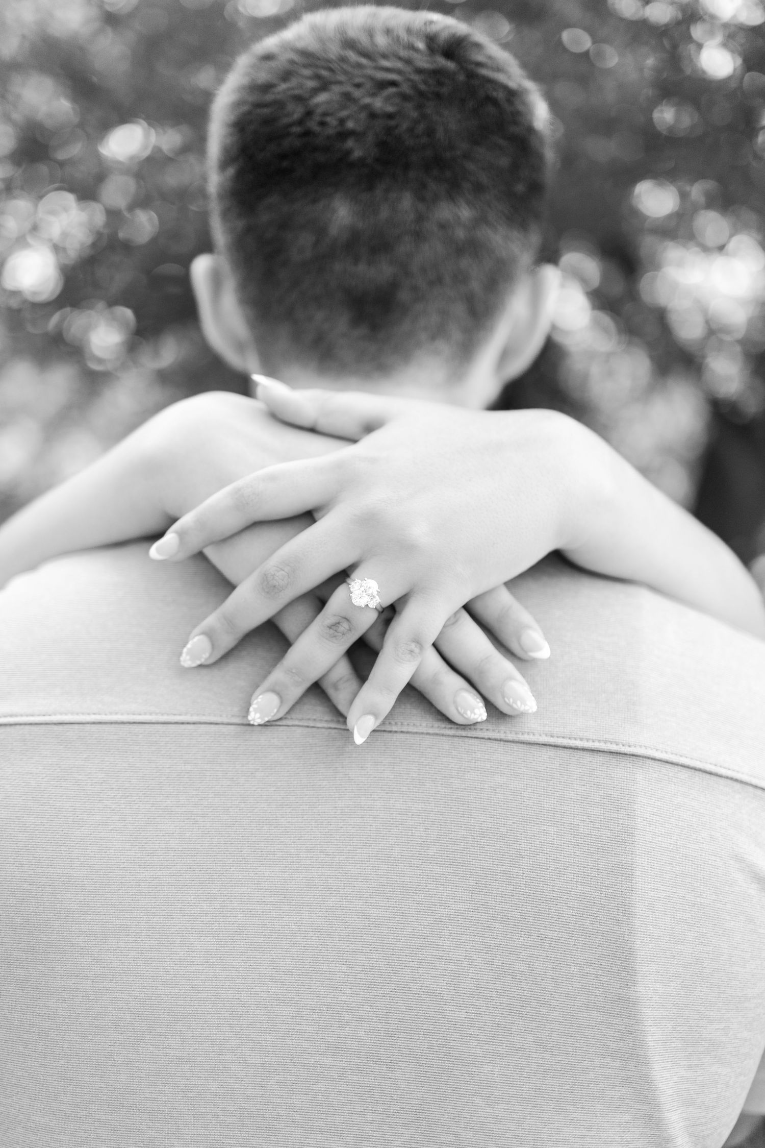 Black and white photo of a woman's hand with engagement ring resting on a man's back during a romantic embrace.