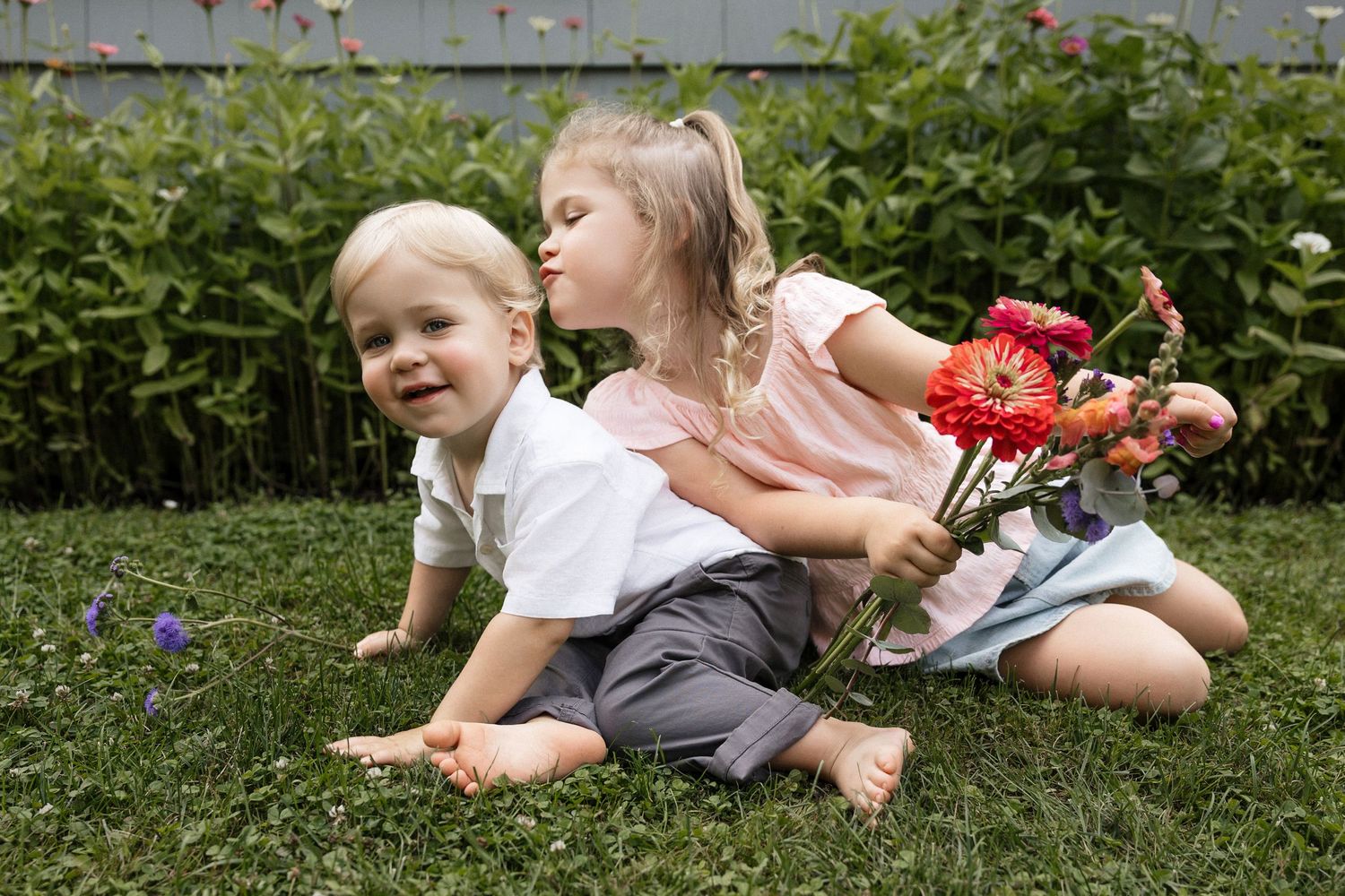 Two young children sitting on grass outdoors, girl kissing boy on cheek while holding colorful flowers.