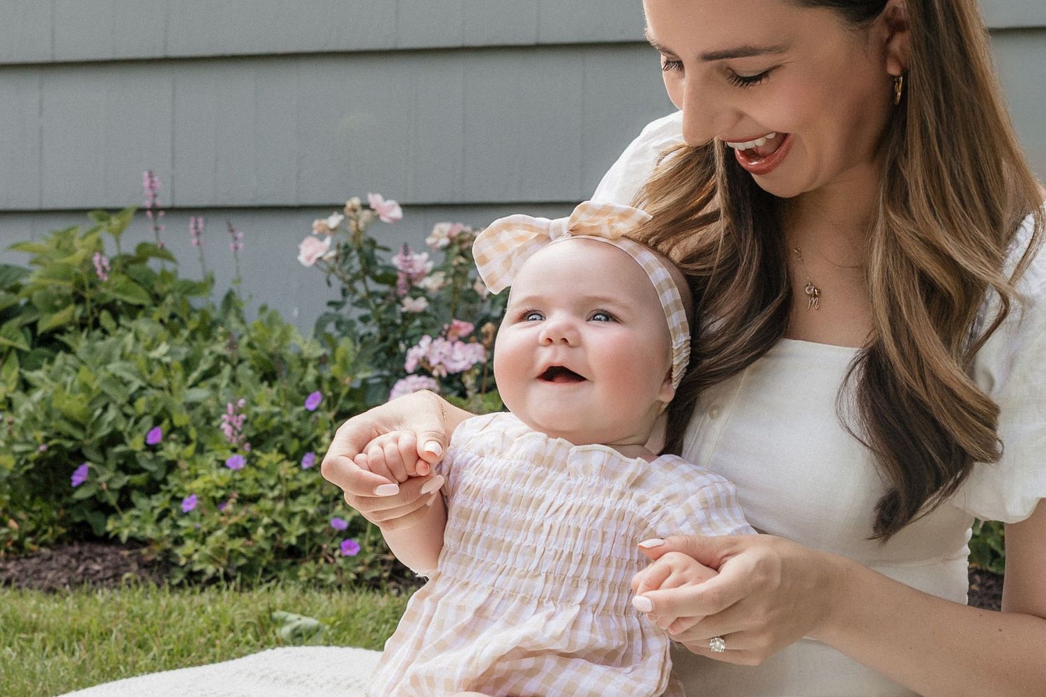 A smiling mother holds her happy baby girl outdoors near a garden, both wearing light pink and white outfits.