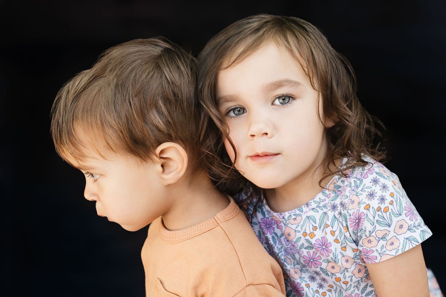 Two young children with brown hair pose together against a dark background, the older girl resting her chin on the younger child.