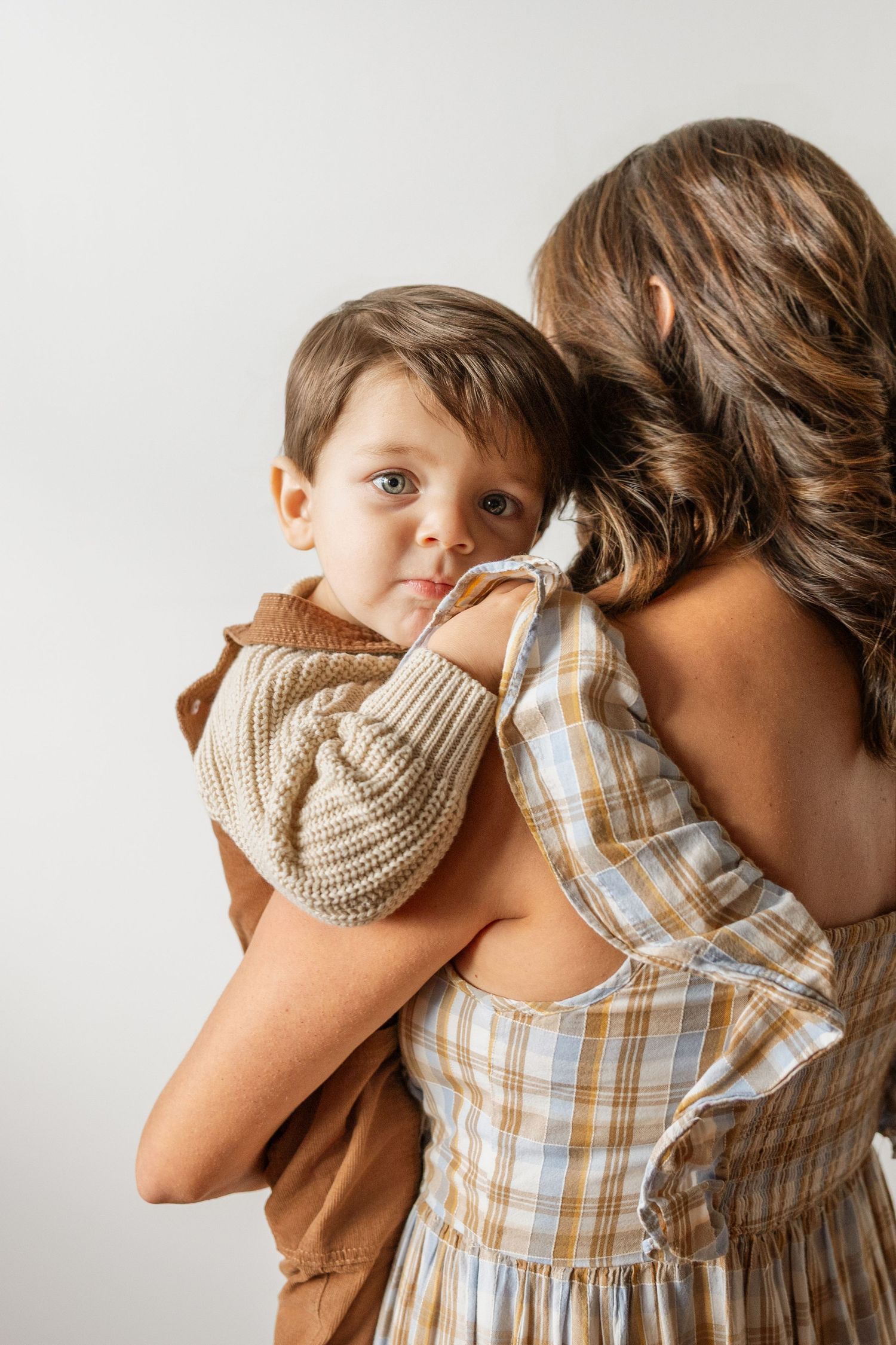 Mother in plaid dress holding young boy in brown outfit against white background.